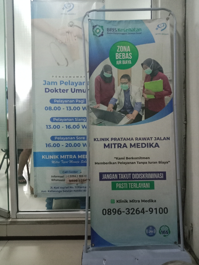The image displays an indoor or semi-indoor area of a medical facility. In the foreground, a freestanding A-frame banner presents information for "KLINIK PRATAMA RAWAT JALAN MITRA MEDIKA" (Mitra Medika Primary Outpatient Clinic). The banner features a graphic depicting three masked medical personnel—one male doctor seated with a laptop, flanked by two female staff members. Accompanying text highlights "ZONA BEBAS IUR BIAYA" (Free Fee Zone), a commitment to free services, and an anti-discrimination policy, along with a contact number (0896-3264-9100) and BPJS Kesehatan and Mitra Medika logos.
Behind a clear glass panel to the left, a second white poster provides "PENGUMUMAN Jam Pelayanan Dokter Umum" (General Practitioner Service Hours) spanning 08:00-13:00, 13:00-16:00, and 16:00-20:00. This poster also identifies the "KLINIK MITRA MEDIKA" and lists its address as Jl. Kyai Asy'ari No. 11 Plantaran, Kaliwungu Selatan, Kendal, Indonesia. Through the glass, the lower legs and feet, wearing sandals, of a seated individual are partially visible. The immediate environment consists of a light-colored floor and walls, with reflections on the glass panel.