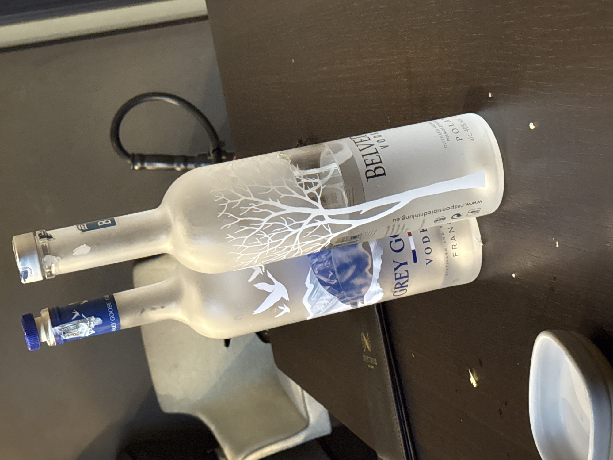 The image depicts two partially full bottles of vodka on a dark brown table. The bottle on the left is a Grey Goose bottle with a blue cap. The bottle on the right is a Belvedere Vodka bottle with a silver cap and a frosted white design of a tree. A small white dish is visible in the lower left corner of the image. The background appears to be an indoor setting with a grey wall and a blurred chair. There are no people or discernible actions taking place. The lighting suggests an indoor environment, possibly a restaurant or bar. The location is not specified to be Sofia, Bulgaria.