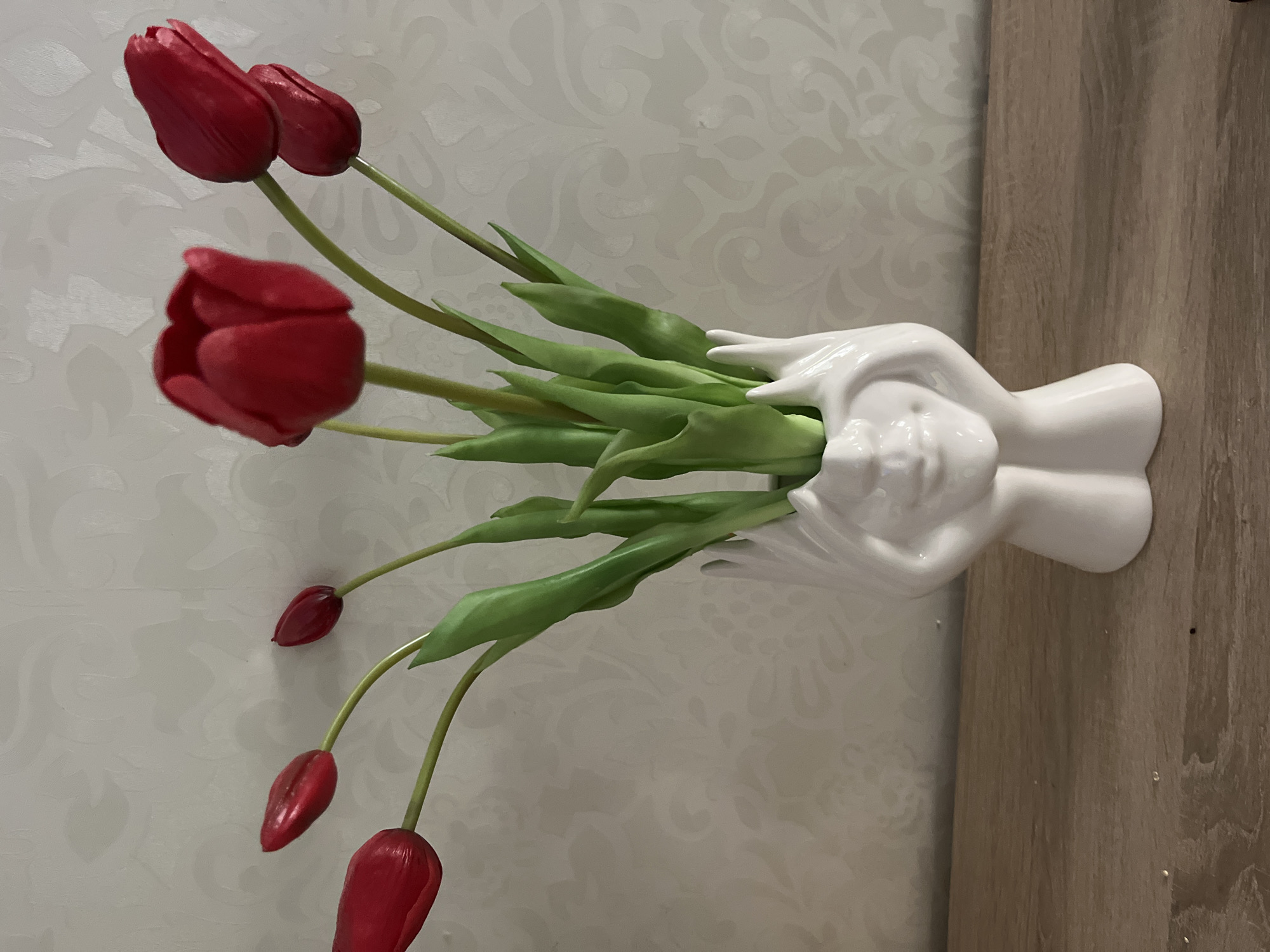 This is a medium shot of a white ceramic vase shaped like a face with hands covering its ears. The vase holds a bouquet of red artificial tulips with green stems and leaves. The vase is placed on a wooden shelf. The background is a light-colored wall with a subtle embossed pattern. The image was taken indoors, likely in a domestic setting. There is no indication of the city of Kharkiv, Ukraine, in the image.