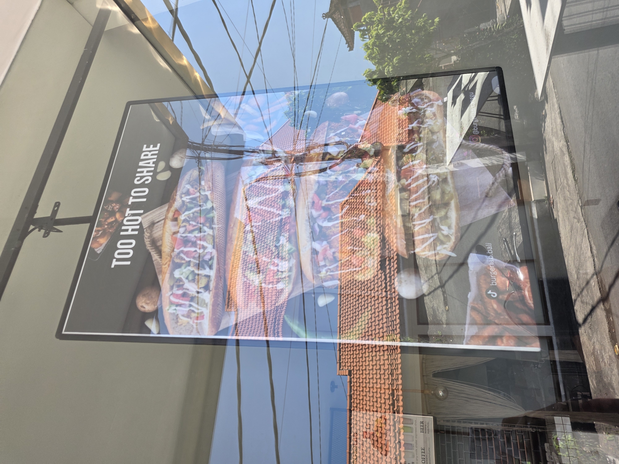 The image shows a poster in a window, likely advertising a restaurant. The poster displays several sandwiches, possibly subs or hot dogs, with various toppings. The text "TOO HOT TO SHARE" is visible on the poster, along with social media handles "@burgerlab.canggu" and "burgerlab.bali," suggesting a restaurant named "Burger Lab." Reflections in the window show the surroundings, including a building with a red tiled roof, power lines, vegetation, and a menu board with "COFFEE" and "BEER" visible, indicating a cafe or restaurant. Based on the social media handle, the location is likely Canggu, North Kuta, Indonesia.