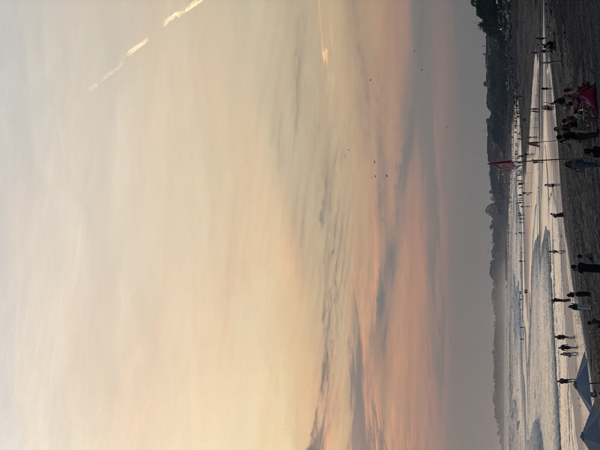This is a photograph of a crowded beach during sunset in North Kuta, Indonesia. The sky is a gradient of pale orange and pink hues, with wispy clouds and a faint contrail from an airplane visible in the upper right quadrant. The ocean is calm, with gentle waves lapping at the shore. The beach is populated with numerous people, some walking along the water's edge, others sitting or standing in groups. There are several beach umbrellas and a few red flags planted in the sand. The background shows a treeline and buildings, suggesting a developed coastal area. The overall mood is relaxed and serene, characteristic of a tropical sunset.