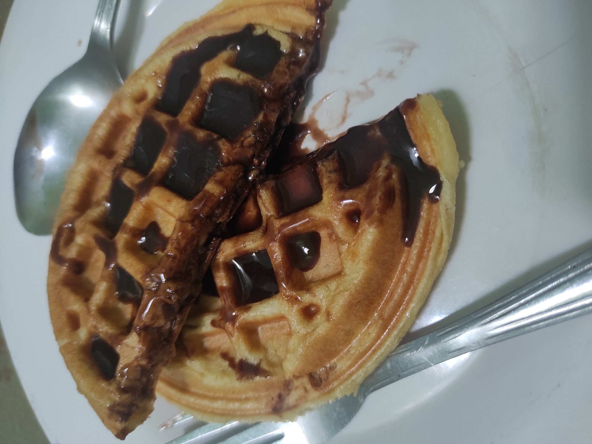 Two halved waffles are stacked on a white plate, drizzled with chocolate sauce. A fork is positioned to the left of the waffles, and a spoon is to the upper right. The background is a light-colored surface, likely a table. The scene depicts a close-up of a dessert.
