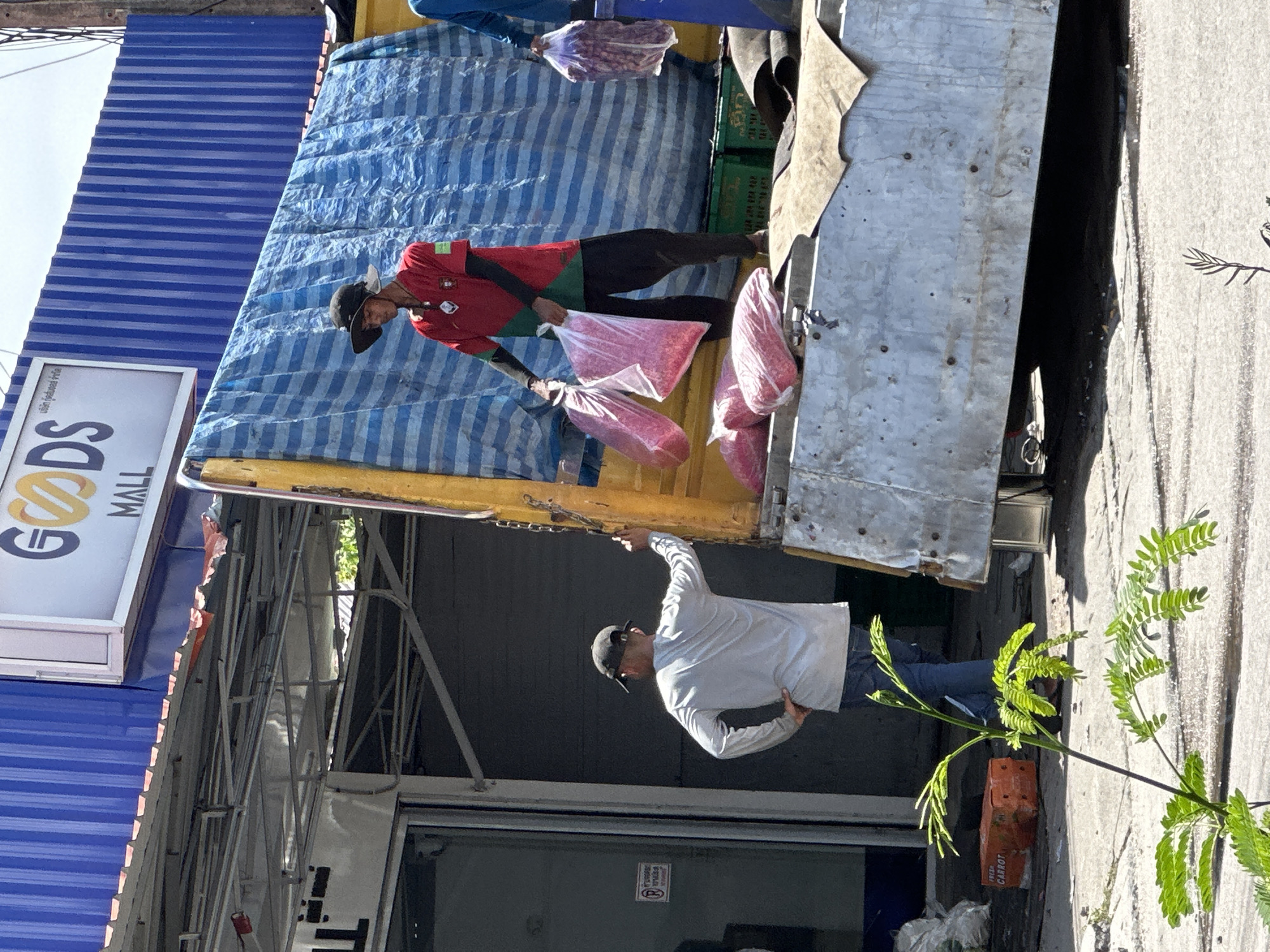 In ตำบลเกาะพะงัน, Thailand, a man in a red Portugal jersey and dark pants hands a bag of red items from the back of a truck to another man on the ground. The truck has a blue and white striped tarp. Next to the truck is a storefront with a sign that says "GOODS MALL".