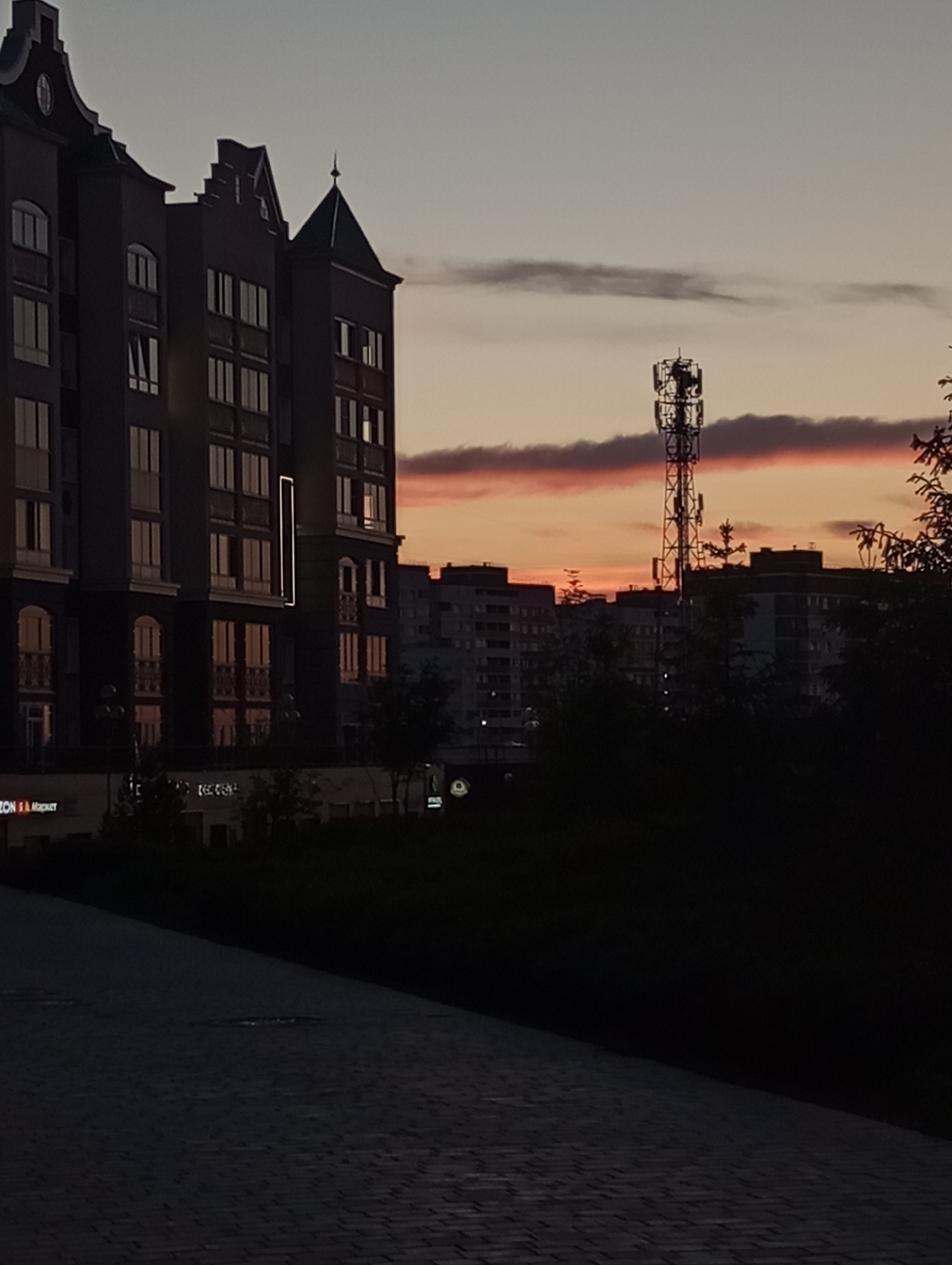 The image shows a cityscape at dusk in Pervomaysky, Russia. A modern, multi-story building with distinctive Dutch-style gabled roofs is prominently featured on the left. Its windows reflect the colors of the setting sun. Further back, a cell phone tower is visible against the orange and grey sky. Other buildings, partially obscured by trees, stretch across the middle ground. A paved road or walkway dominates the foreground and is partially in shadow. There are no people visible in the image.