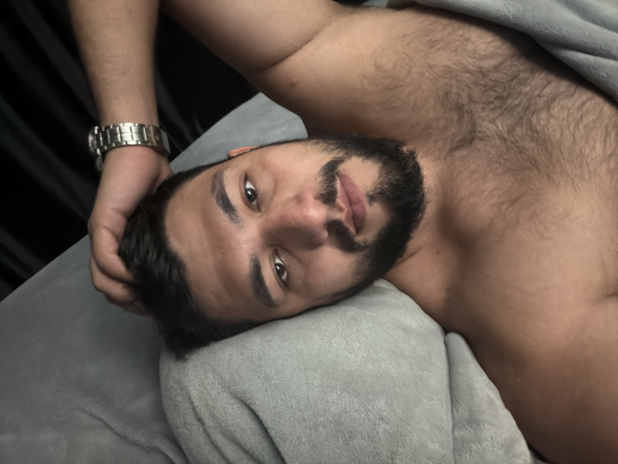 A man with dark hair and a beard is lying on a grey pillow, looking at the camera. He is shirtless and has a silver wristwatch on his left wrist. A grey blanket is partially covering his upper body. A dark curtain is visible in the background. No information is provided to determine the location.
