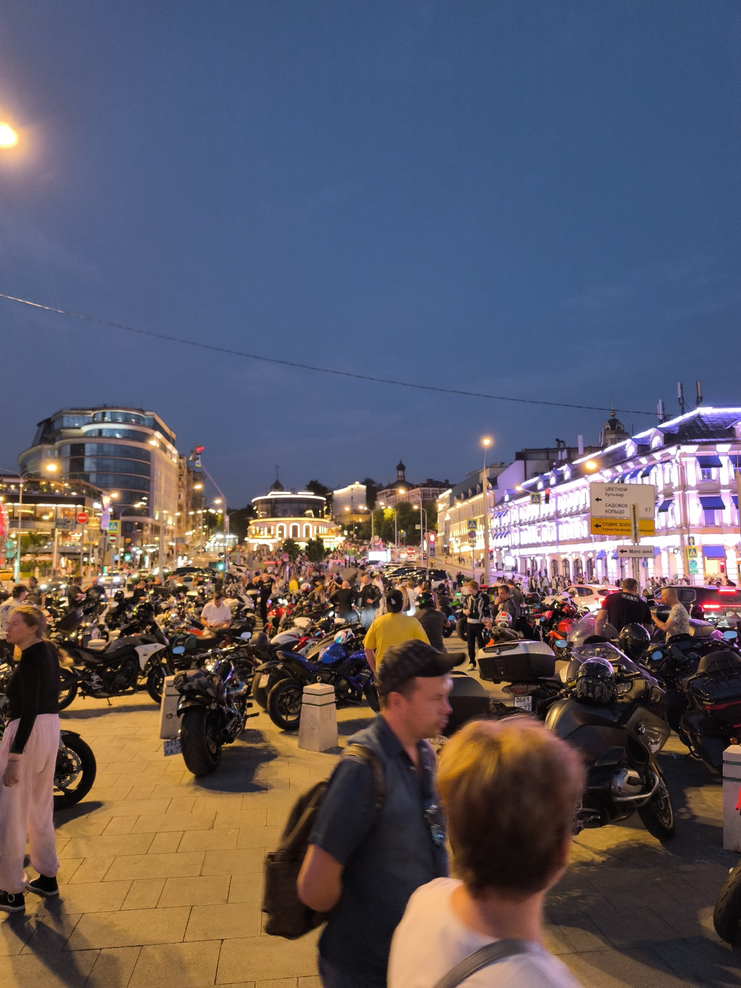 The image captures a bustling street scene in Moscow, Russia, likely during an evening event. A large gathering of motorcycles is parked on a paved area, with many people milling around. The background features illuminated buildings with modern and classical architectural elements, characteristic of a city center. Streetlights and building lights cast a warm glow on the scene, while the sky is a deep twilight blue. Signs in Russian are visible, one indicating directions to "Tsvetnoy Boulevard" and "Sadovoye Koltso," a major ring road in Moscow. The atmosphere appears lively and social, with individuals dressed in casual attire. Some people are walking, others are standing and talking, and a few are near the motorcycles, suggesting a possible motorcycle rally or gathering. The overall impression is one of urban activity and community engagement.