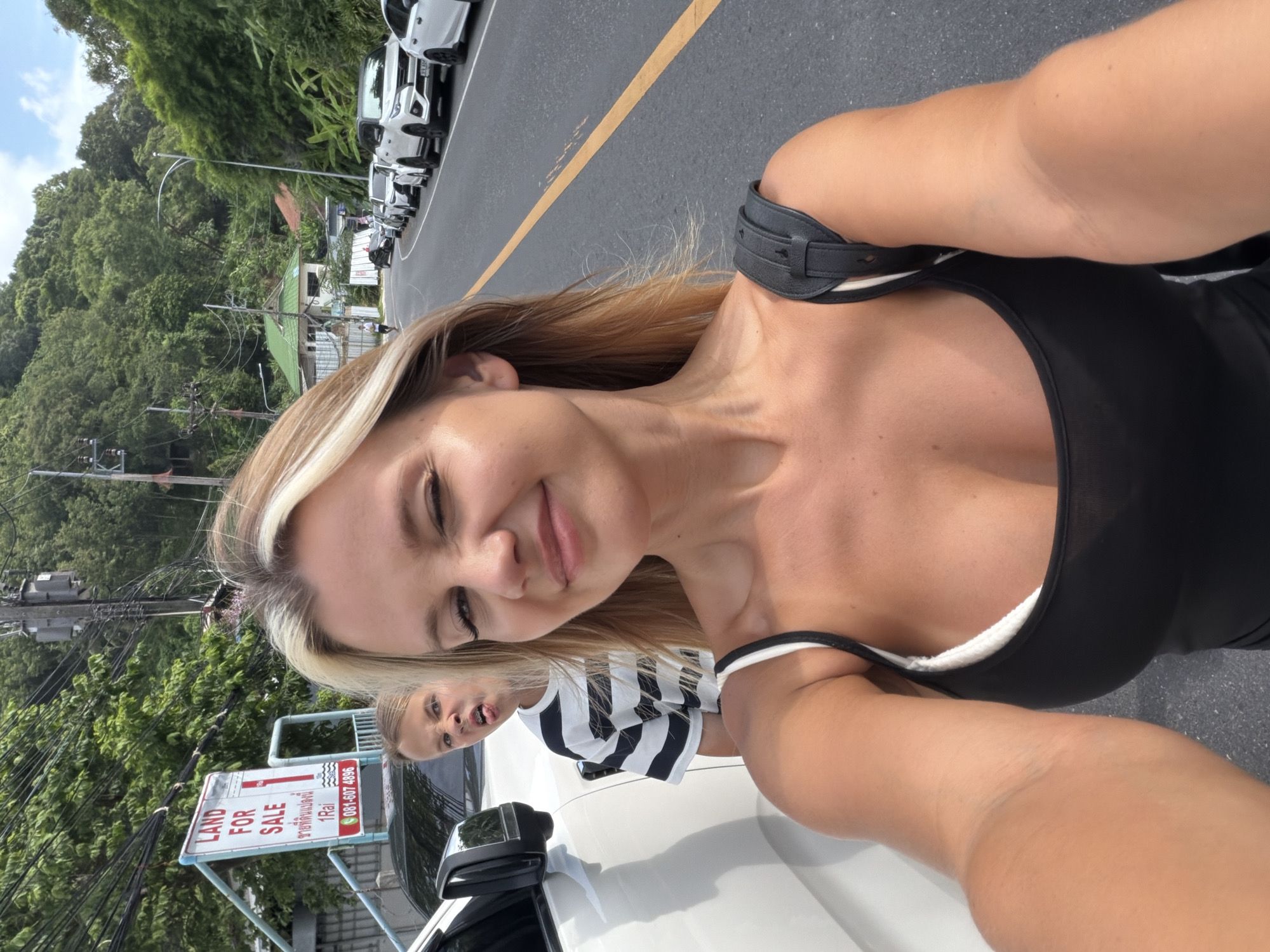 A young woman with blonde hair is taking a selfie on a sunny day, standing next to a white car. She is wearing a black tank top and winks at the camera. Behind her, a young boy with blonde hair makes a funny face, sticking his tongue out. A "Land for Sale" sign is visible behind them, with Thai script and a phone number. They appear to be on a street in Wichit, Thailand, with a line of cars parked along the road and lush green hills in the background. The scene suggests a casual, possibly vacation-related moment captured outdoors.