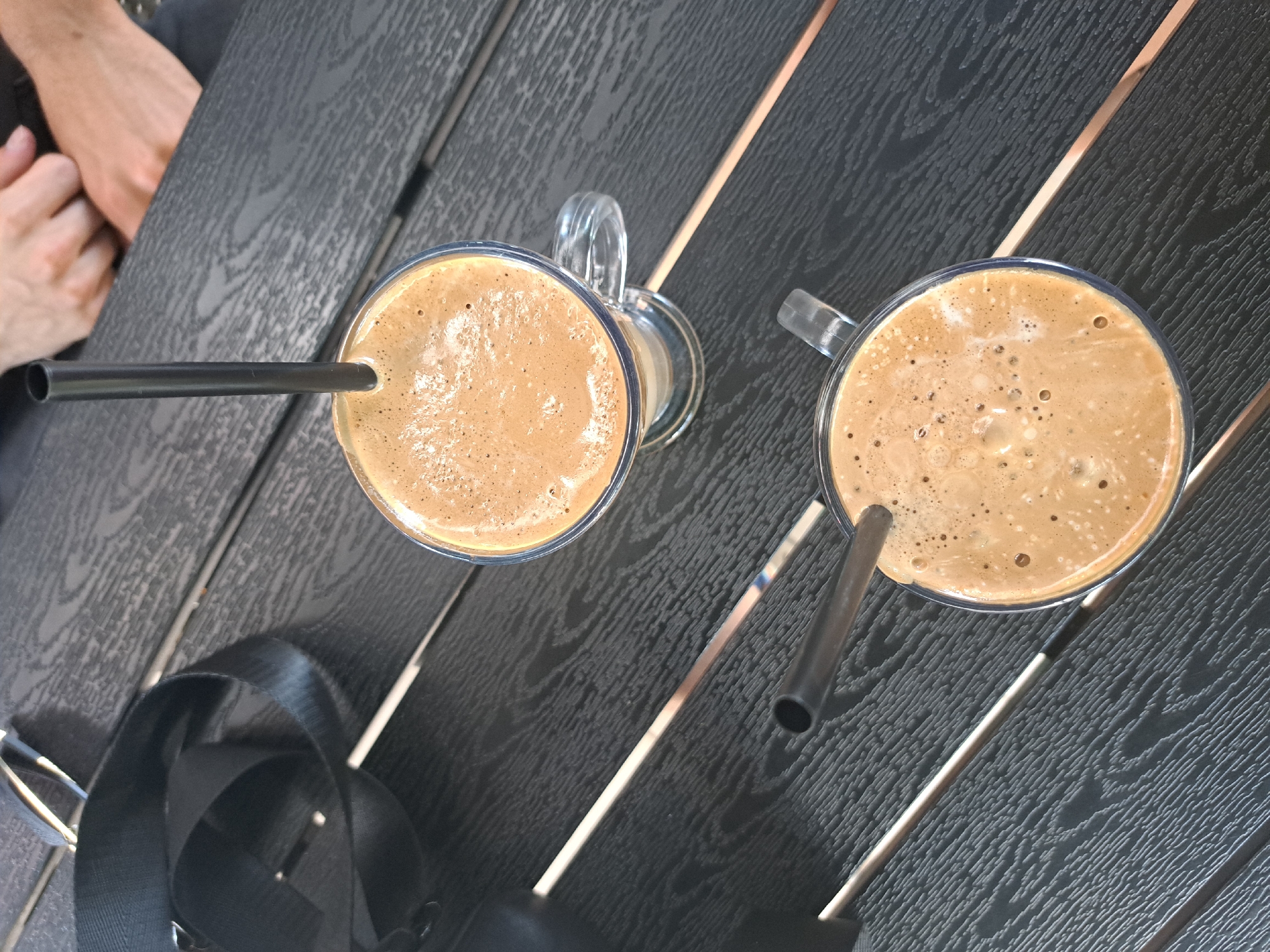Two clear glasses filled with a frothy, light brown beverage, possibly coffee or a milkshake, are placed on a dark wooden table. Each glass has a handle and contains a dark straw. One straw extends diagonally to the left, entering the left glass. The other straw is positioned on the right side, entering the right glass and extending downwards. The table surface has a wood grain texture with a pattern of alternating dark and lighter strips. In the upper left corner of the image, a portion of two people's arms and hands are visible, suggesting they are sitting at the table. A black strap, possibly from a bag or camera, is draped across the lower left section of the table. The lighting appears to be natural, indicating it could be daytime. The scene suggests a casual setting, perhaps an outdoor cafe or a relaxed gathering. No visible text is present in the image.