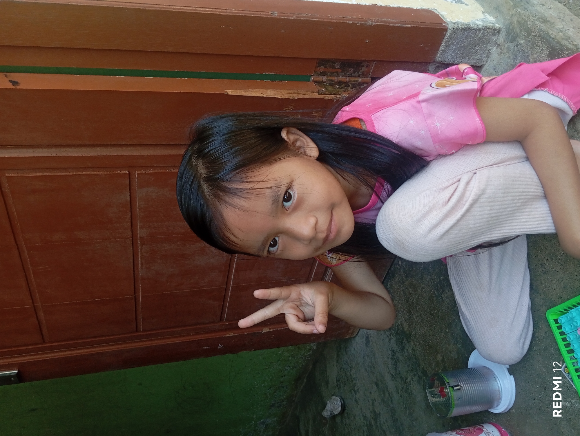 A young girl with dark hair and brown eyes is sitting on a concrete floor in front of a wooden door. She is wearing a pink t-shirt and light-colored pants. Her right hand is raised, and she is holding up two fingers in a peace sign. A gray stone is on the floor to her left, and a silver can with a white base is next to it. To her right, a green basket with blue and pink objects is visible. The background includes a green wall and a brown wooden door with visible hinges. The scene appears to be an outdoor setting, possibly a residential area in Pagar Alam, Indonesia.