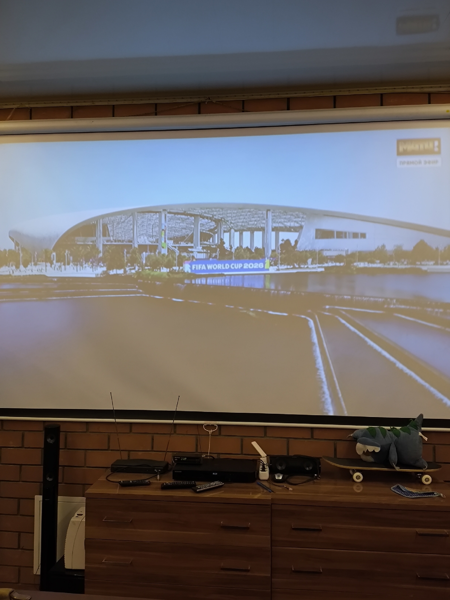 In the city of Pokrov, Russia, an image captures a scene focused on a projection screen displaying a broadcast of the FIFA World Cup 2026. The screen is illuminated, showing a modern stadium structure with an expansive roof. In the lower portion of the frame, a wooden dresser is visible, adorned with various electronic devices, including a soundbar, DVD player, and remote controls. A plush shark toy is positioned on a skateboard atop the dresser. Two television antennas are also present, along with a tall black speaker. The backdrop is a brick wall, indicating an indoor setting, likely a home entertainment area.