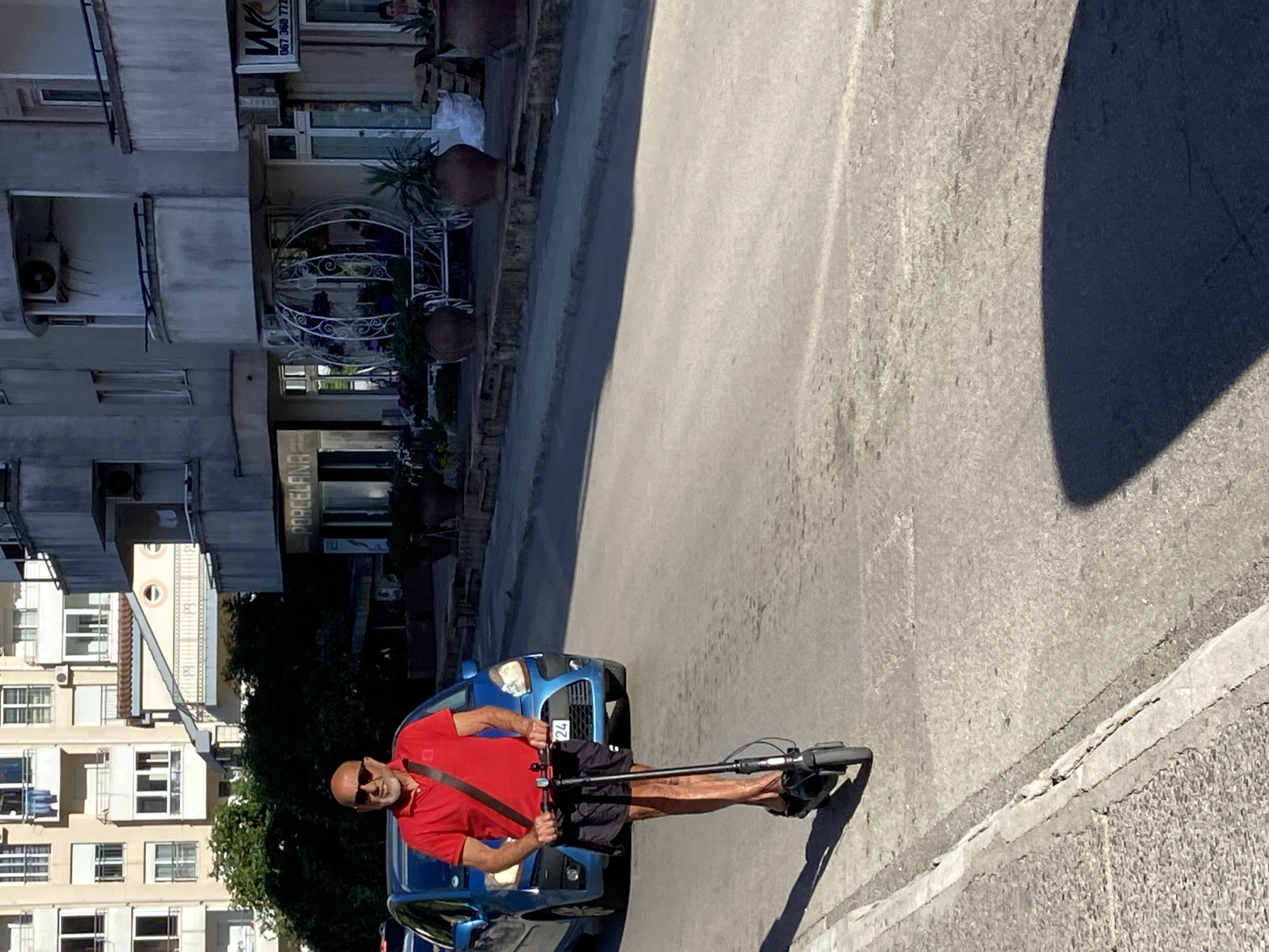 An outdoor scene in Budva, Montenegro, on a clear, sunny day shows a bald, bearded man, appearing middle-aged to senior, riding a black electric scooter on an asphalt road. He wears a red short-sleeved polo shirt, dark shorts, sunglasses, and a dark cross-body bag, and smiles towards the viewer. Behind him, a blue minivan is visible with a partially discernible license plate featuring "24".
To the left of the road, multi-story residential buildings with light-colored facades and numerous windows and balconies line the street; laundry is visible hanging on one distant balcony. Green foliage partially obscures the lower parts of these buildings. To the right, a row of commercial ground-floor establishments includes a shop labeled "PORCELANA," another featuring a large white, ornate, cage-like structure adorned with potted plants, and a third with a distinct "W" logo and a telephone number (067 360 772X). Several large potted plants are positioned along the pavement in front of these shops. A concrete curb runs along the left edge of the road. Strong sunlight from an unseen source casts sharp, dark shadows across the street surface.