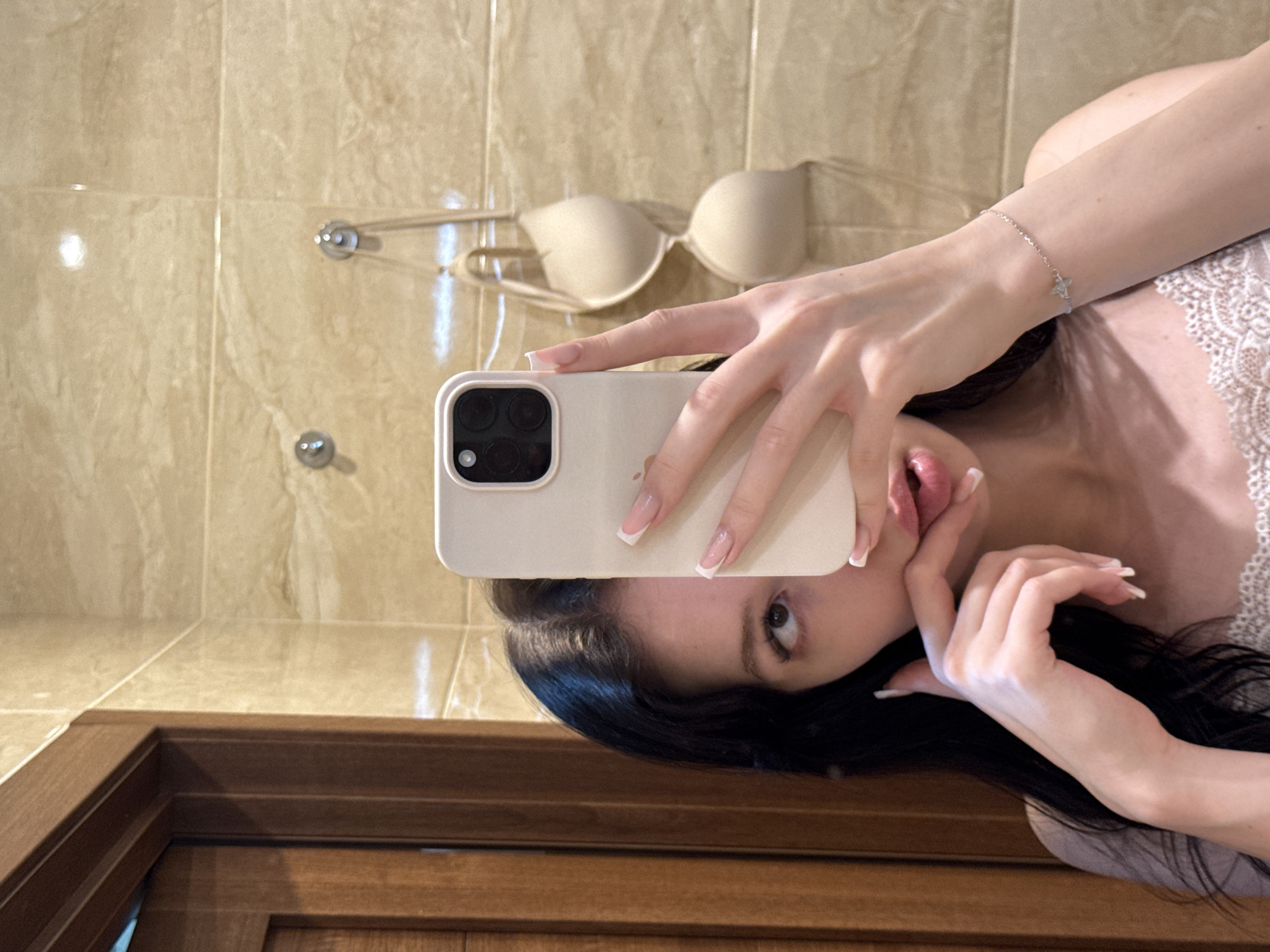 A young woman with dark hair is taking a mirror selfie in a bathroom with beige tiled walls. She is holding a beige iPhone in her right hand, partially obscuring her face. She is wearing a white lace camisole and a delicate silver bracelet with a small charm. A beige bra is hanging on hooks on the wall behind her. To the left of the frame is a wooden door. The image appears to have been taken in a residential bathroom. The city of Khimki, Russia is not explicitly indicated by any visual cues in the image itself.