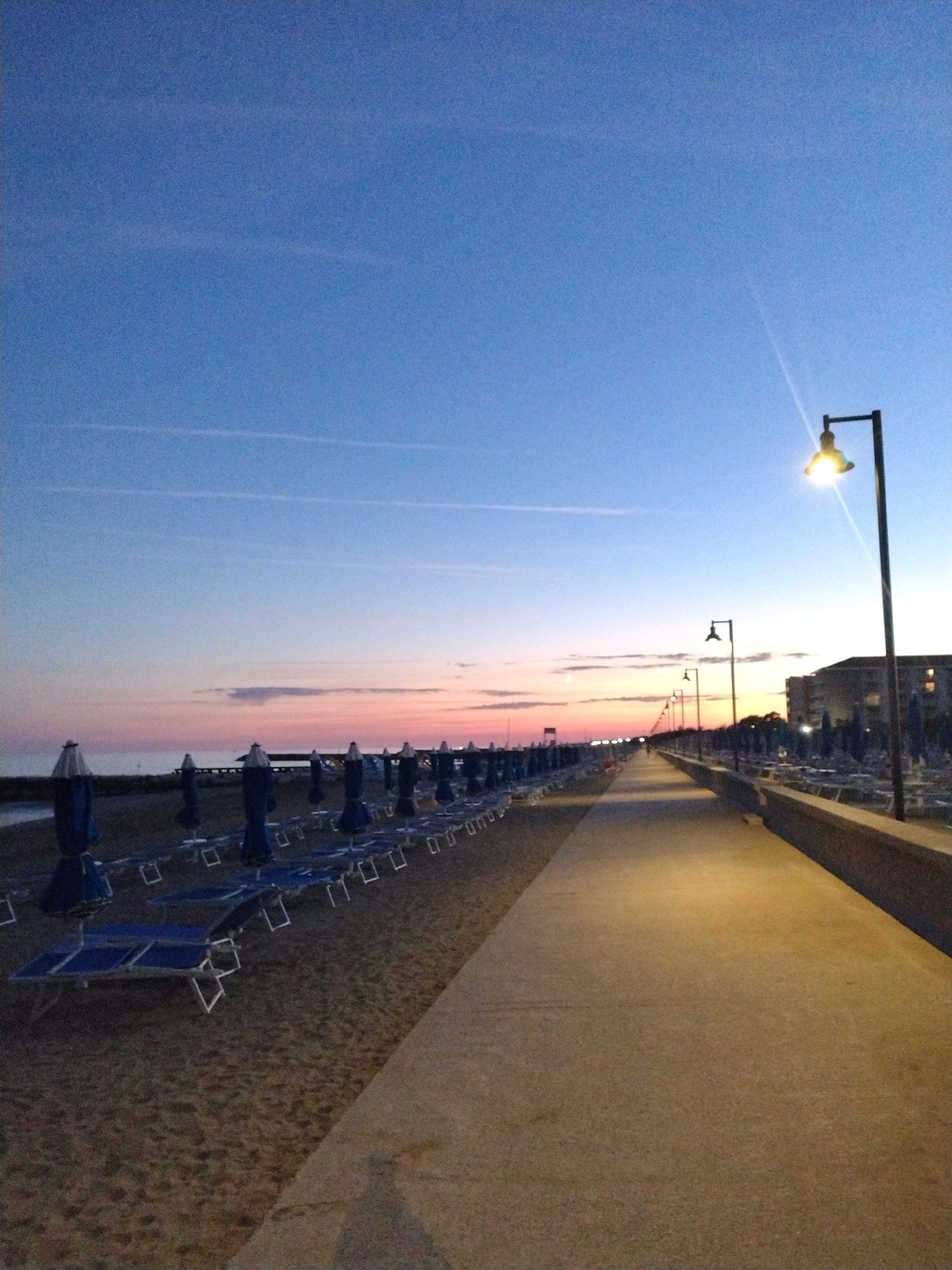 The image shows a beach promenade at dusk in Caorle, Italy. The sky transitions from a deep blue at the top to hues of pink and orange near the horizon. Several streetlights are illuminated along the promenade, casting a warm glow. Rows of closed beach umbrellas and lounge chairs are neatly arranged on the sandy beach to the left of the promenade. In the background, buildings can be seen. The promenade itself is a paved walkway, appearing empty of people. The scene evokes a sense of tranquility following a day at the beach.