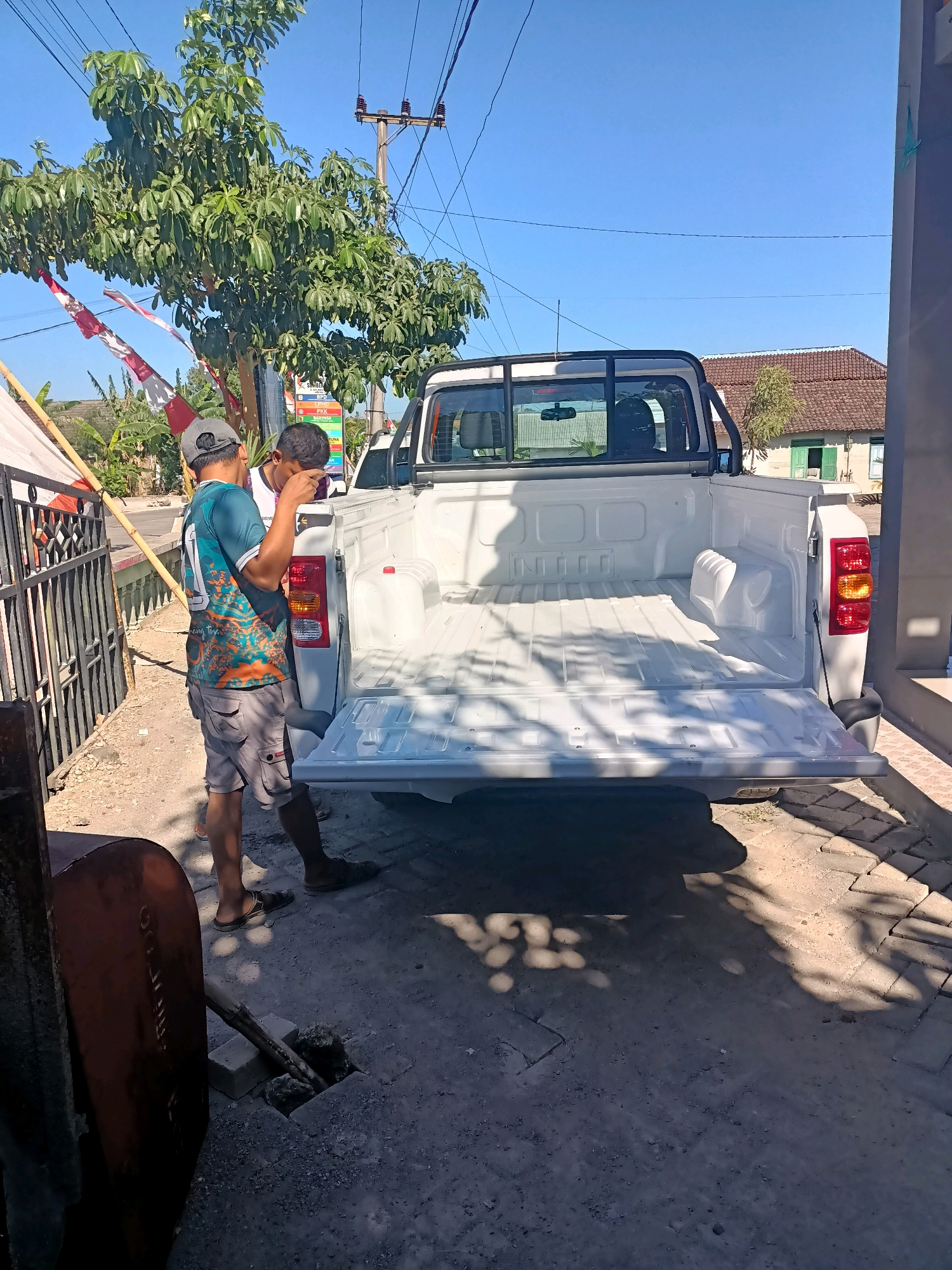 This image depicts a scene in Sumber Bendo, Indonesia, where a white pickup truck is parked. Two individuals are near the rear of the truck. One person, wearing a teal shirt with an orange design and a grey cap, is standing and looking towards the truck bed. The other person, with dark hair and wearing a dark shirt, is standing beside the first person, with their head down as if looking at something on the truck. The truck bed is open and appears to be empty. The environment shows a dirt and gravel ground surface, with some patchy vegetation and paving stones. In the background, there is a tree with red and white Indonesian flags attached to it, a utility pole with wires, and a building with a tiled roof and green shutters. The sky is clear and blue, suggesting a sunny day.