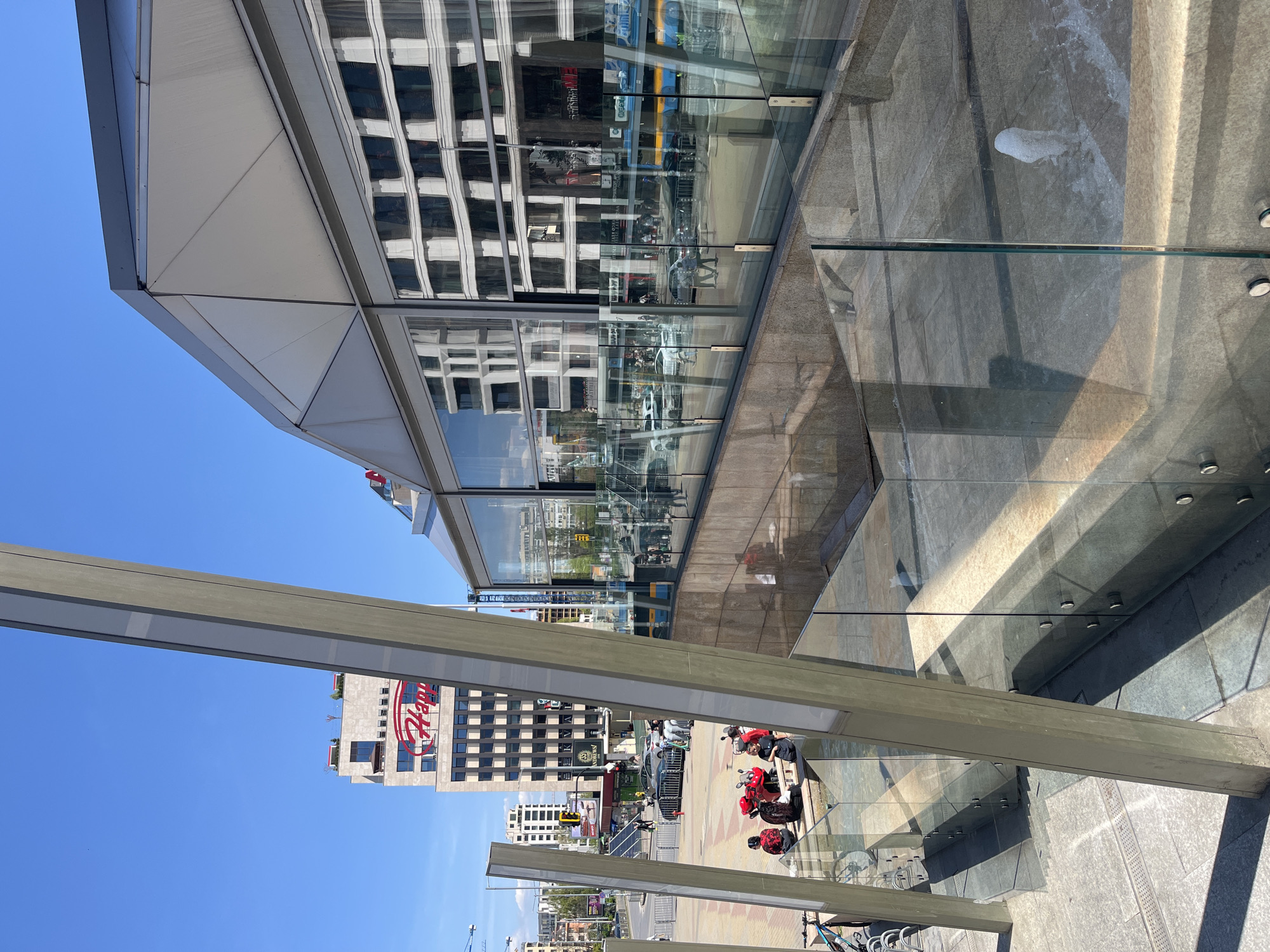 A street-level view from Sofia, Bulgaria. The image shows modern architecture and street life. A tall building with a glass facade reflects other buildings and the sky. A building with the "Happy" logo is visible in the background. There are several red scooters parked on the pavement along with some bicycles, and people are present on the sidewalks. The sky is blue and clear. The image seems to be taken during the day.