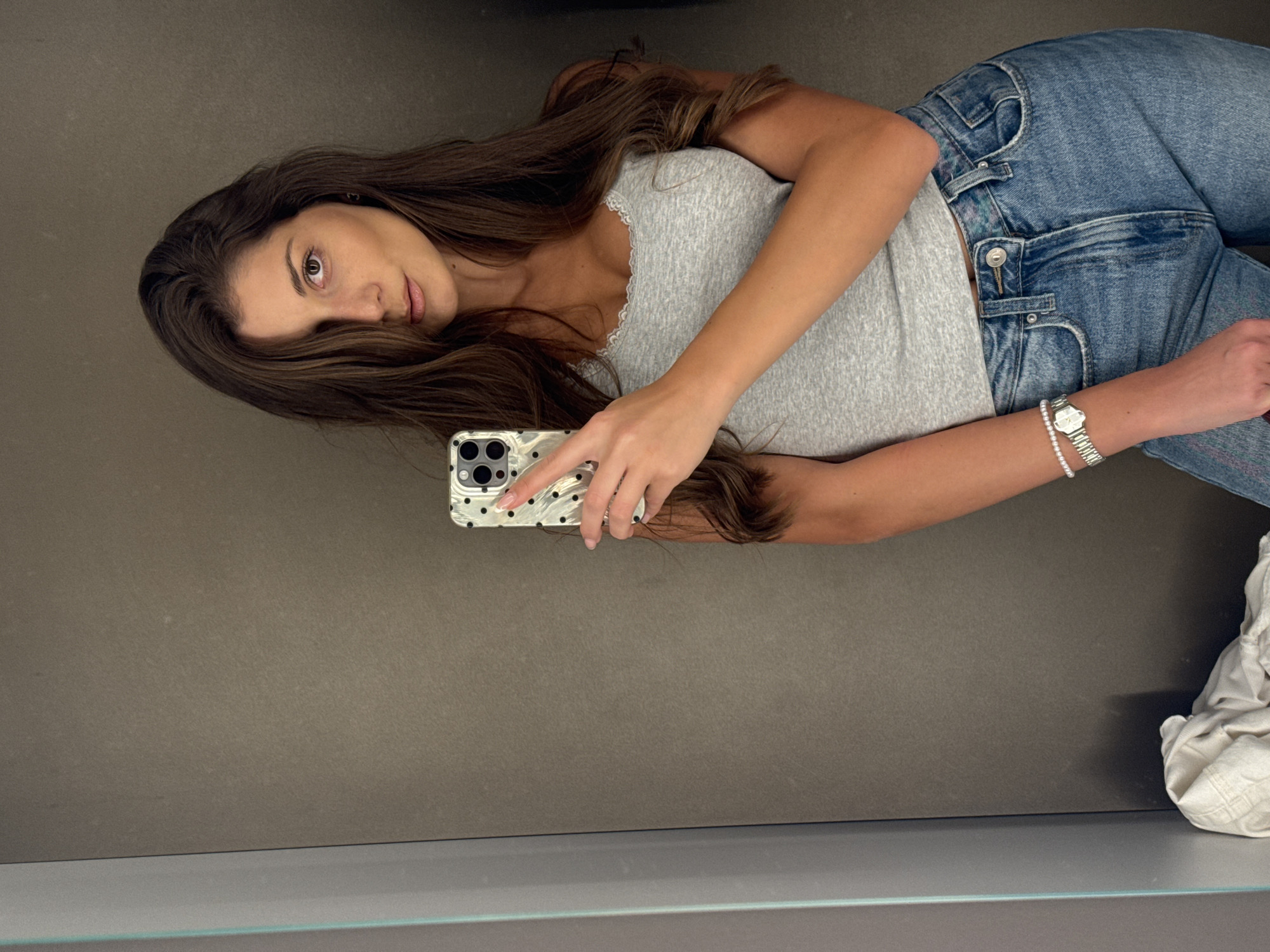 A young woman is taking a selfie in what appears to be a clothing store fitting room. She is holding her phone in her right hand, which is angled slightly towards the mirror. She is wearing a grey tank top, blue jeans, and a watch and bracelet on her left wrist. Her hair is long and brown. The background is a simple grey wall, and there is a shelf at the bottom of the frame, with a pile of white clothes sitting on it. The image provides no data related to Burgas, Bulgaria.