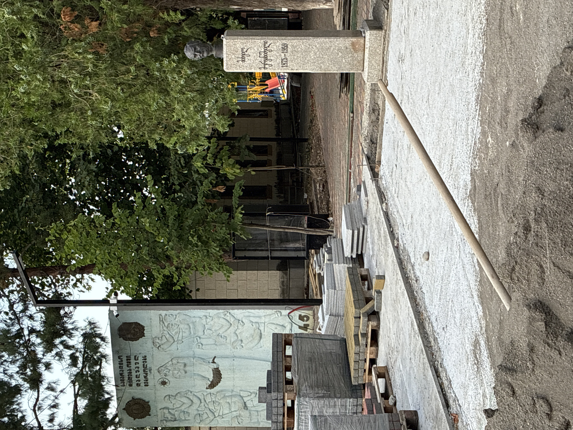 In Surami, Georgia, a public area is undergoing construction. Piles of grey and red pavers are stacked on wooden pallets in the foreground, indicating ongoing landscaping or street work. To the right, a granite monument features a bust of a man and inscriptions in Georgian script, with the dates "1875-1959" visible. Behind the monument, a large, light-colored mural or relief depicts figures in a historical or commemorative scene. In the background, trees with lush green foliage and a brick building are present, along with a glimpse of a colorful children's playground. The ground surface shows areas of gravel and a thin layer of white material, suggesting preparation for paving. A wooden-handled tool lies across the prepared ground. The sky is overcast.