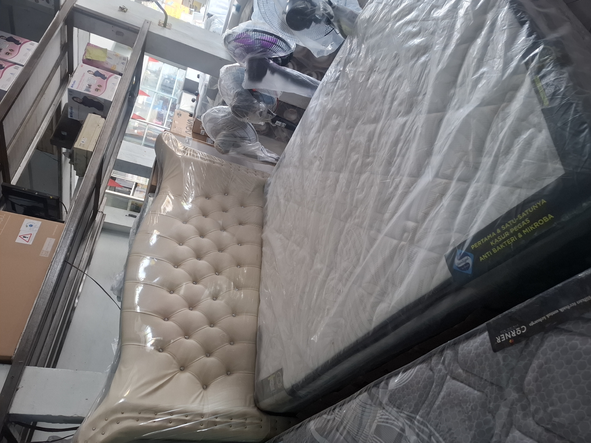 The image shows a store that sells mattresses and fans. In the foreground, a bed with a cream-colored, tufted headboard is covered in plastic wrap, with a mattress also covered in plastic. To the left, another mattress is partially visible, also wrapped in plastic. In the background, several oscillating fans are on display, some of which are also covered in plastic. Shelves in the background are stocked with various items, including boxes and electronics. The lighting suggests an indoor environment, possibly a showroom or warehouse. The general impression is that of a retail space. Based on the provided location, this scene is taking place in a store in Banjarmasin, Indonesia.