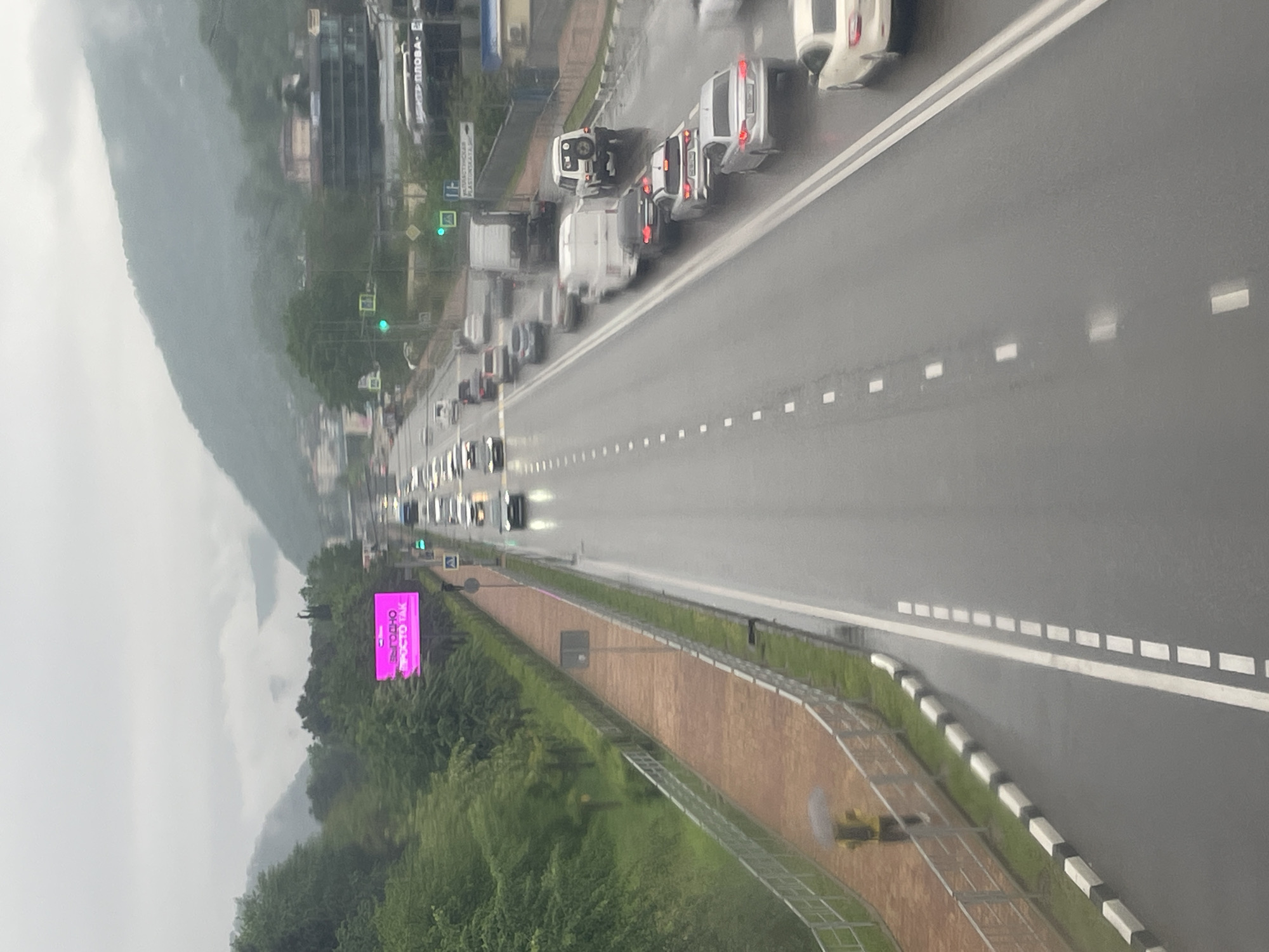 The image depicts a wet, overcast day in Sochi, Russia, showing a multi-lane highway with heavy traffic moving in one direction. A billboard is visible next to a wooded area adjacent to the road. Distant buildings and a large mountain slope are partially obscured by fog or rain. The road markings and median strip are visible, along with a pedestrian walkway protected by a metal guardrail. The overall atmosphere is one of a busy city highway experiencing inclement weather.