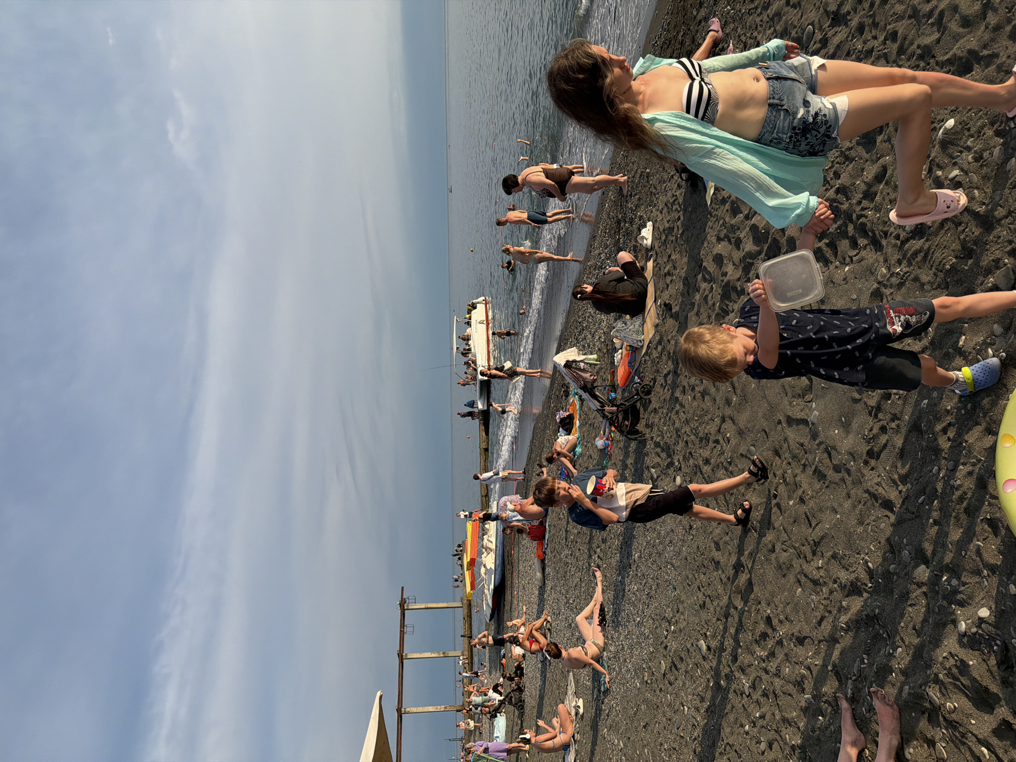 This is a photograph taken on a beach in Sochi, Russia, likely during the daytime with clear weather. The scene depicts a bustling beach with many people enjoying various activities. The foreground is dominated by dark, pebbly sand, with some people seated or lying on it. A young boy in a dark t-shirt and shorts is holding a transparent plastic container, looking intently at something in his hand. To his left, a woman in a striped bikini top and denim shorts is reclining on the sand, wearing a light blue cover-up and pink slippers.
Further back, along the water's edge, a group of people are standing or wading in the shallow sea. Some are partially submerged, while others are closer to the shore. A small boat is docked near the shore, with people on and around it. In the background, there are structures that appear to be piers or breakwaters extending into the sea. The overall atmosphere seems lively and relaxed, typical of a summer day at the beach. The sky is a light blue with some wispy clouds, suggesting fair weather. The composition is a mix of close-up shots of individuals and wider views of the crowded beach.