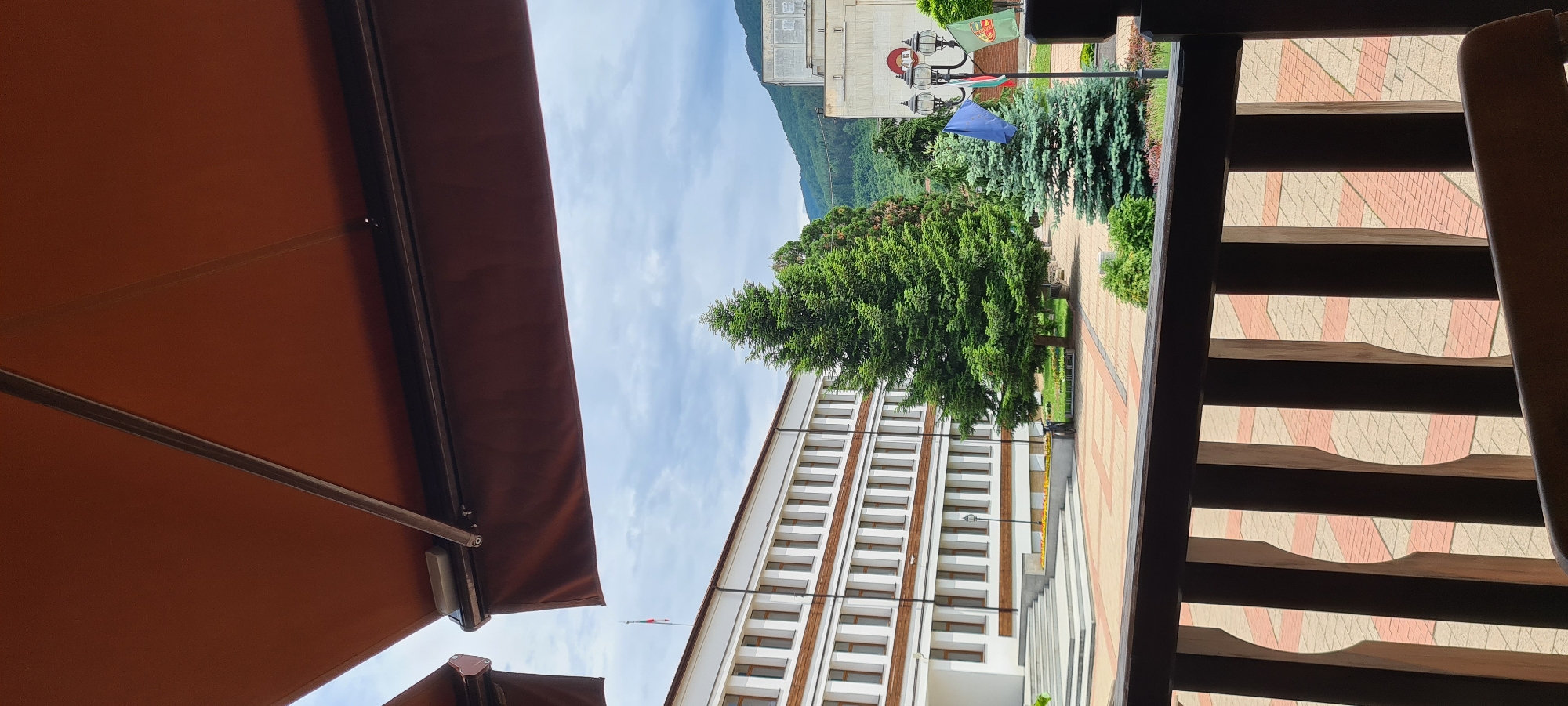The image depicts a public square in Etropole, Bulgaria, viewed from an elevated position behind a dark wooden railing. A reddish-brown retractable awning extends across the upper portion of the frame, partially obscuring the sky.
In the midground, a large, multi-story building with a white facade and numerous rectangular windows with brown frames dominates the left side. A wide, tiered staircase leads to its entrance. Above and behind this building, a Bulgarian flag (white, green, red horizontal stripes) flies from a pole. To the right of this building, a large, dark green coniferous tree stands prominently in the center of a patterned paved square. The square's paving consists of alternating light-colored and reddish-brown rectangular blocks arranged in a geometric design.
Further right, a black, ornate lamppost features multiple light fixtures and three flags: the blue and yellow-starred flag of the European Union, the Bulgarian national flag, and a green flag with an emblem, likely representing the municipality. Adjacent to the lamppost is a smaller, blue-green coniferous tree. Another building, lighter in color, is partially visible on the far right, displaying what appear to be Cyrillic characters ("ДБ") on its upper facade.
The background reveals green, tree-covered mountains rising under an overcast sky filled with white and grey clouds. The wooden railing at the bottom of the frame has vertical balusters and a dark top rail, indicating the vantage point is from a balcony or terrace overlooking the square. No people are visible.