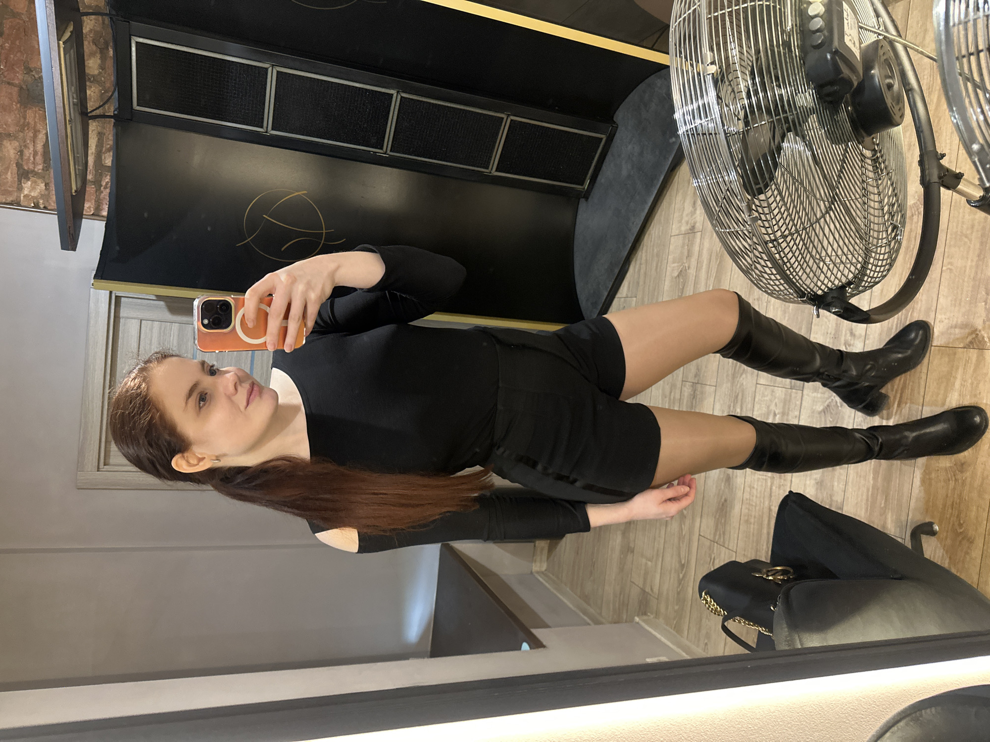 A woman is taking a selfie in front of a mirror. She is wearing a black bodysuit with long sleeves and black shorts, and black knee-high boots. She is holding an orange smartphone.
Behind her are the following items: A black vanity, a brick wall, a gray wall, an industrial fan, a black purse, and a black stool.
There are no indicators as to the location of the image in Riga, Latvia.