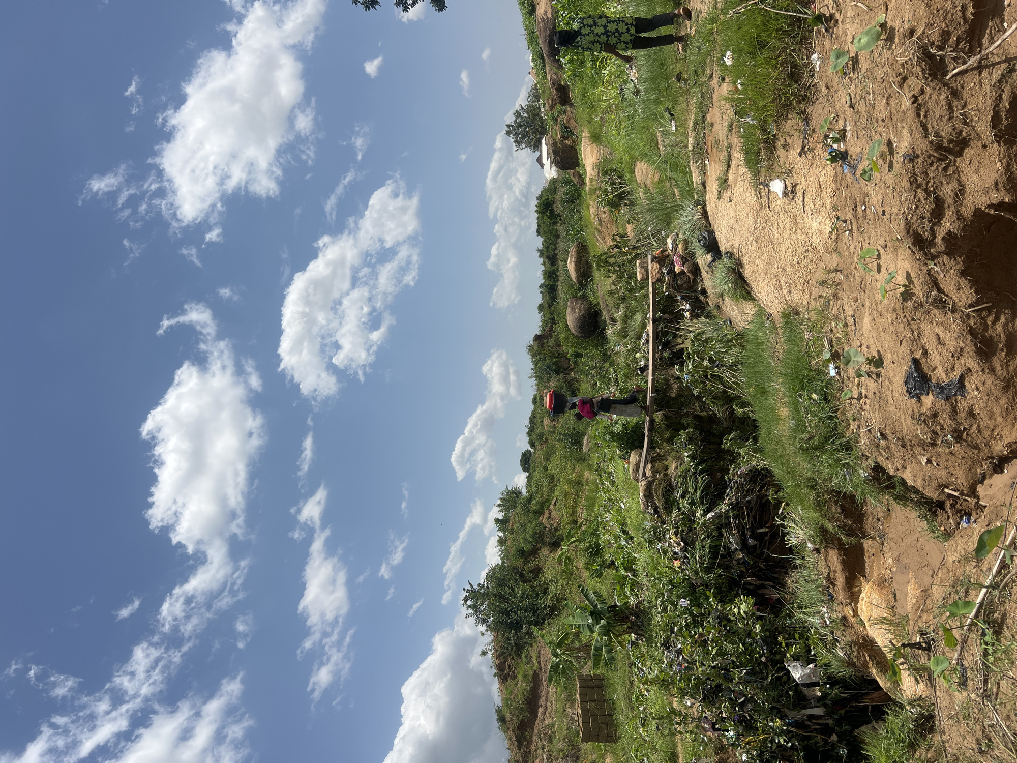 The image depicts a rural landscape with lush green vegetation, dirt paths, and rocky outcrops under a bright blue sky with scattered white clouds. The scene appears to be in or near Jos, Nigeria, judging by the overall environment. In the mid-ground, a person is walking across a narrow wooden bridge over a ditch. This person is carrying a red basin on their head. Further to the right, another person, wearing a green and black patterned shirt and dark pants, walks away from the viewer on a dirt path. The area shows signs of human activity, with piles of bricks stacked on the left, and some scattered debris and plastic waste visible along the ditch and path, suggesting potential environmental challenges in the vicinity. The overall impression is of a functional, perhaps slightly impoverished, rural community.
