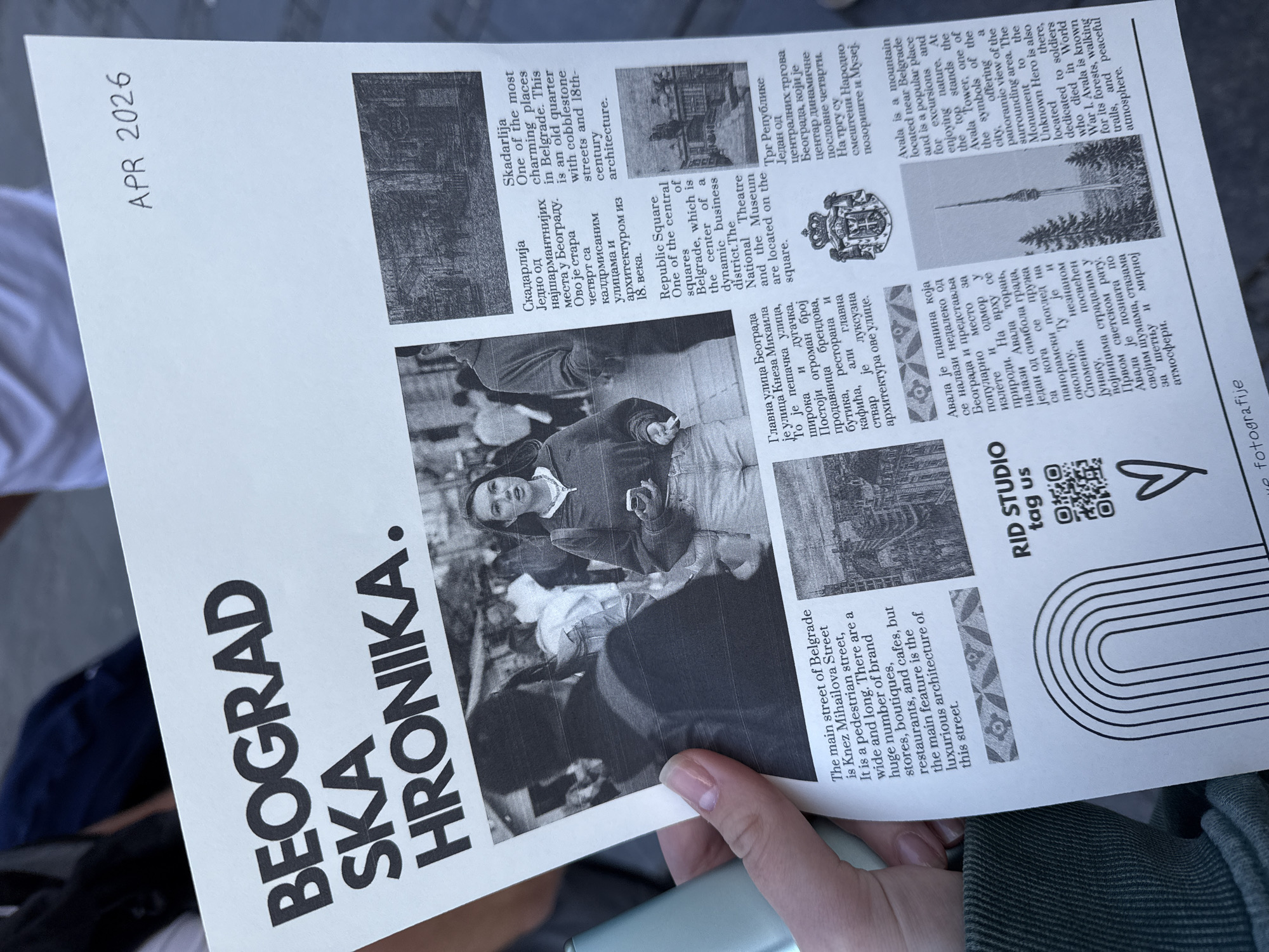 A hand holds a black and white printed document titled "BEOGRAD SKA HRONIKA." (Belgrade Chronicle), with "APR 2026" handwritten in the top left corner. The document serves as an informational guide about prominent locations in Belgrade, Serbia, presented with text in both Serbian and English alongside monochromatic images.
The content highlights several key areas:
* **Skadarlija:** Described as a charming old quarter with cobblestone streets and 18th-century architecture, accompanied by a small image of a historic street.
* **Republic Square:** Noted as a central square, a dynamic business district, and the location of the National Theatre and Museum, with an image of the square.
* **Knez Mihailova Street:** Featured as the main pedestrian street, wide and long, known for its numerous brand stores, boutiques, restaurants, cafes, and luxurious architecture. A large central image shows a person walking on a lively street, likely Knez Mihailova Street.
* **Avala:** Described as a mountain near Belgrade, popular for nature excursions. It features the Avala Tower (a city symbol offering panoramic views) and the Monument to the Unknown Hero (dedicated to WWI soldiers). An image of the Avala Tower is included.
The bottom right of the document displays "RID STUDIO tag us ♡" and a QR code, along with "we fotografije" (likely indicating the source of photography), suggesting it's a publication by this studio. The hand holding the document, partly obscuring some text, is positioned over what appears to be a green garment, with a soft, blurred background, indicating a casual setting.
