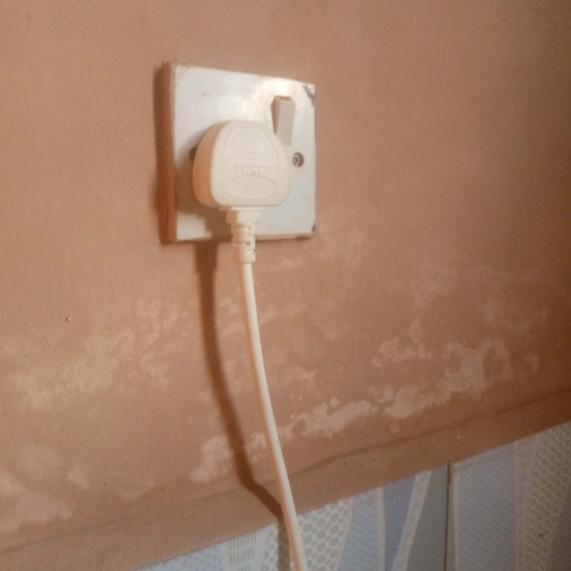 A white three-pin electrical plug with the word "FUSE" embossed on its casing is inserted into a white electrical socket mounted on a light brown wall. The wall has an uneven texture with lighter patches, potentially indicating dampness or discolored plaster. Below the wall is a tiled surface with a pattern of white and light blue geometric shapes. A white electrical cord extends downwards from the plug. The scene is lit from the left, casting a shadow from the plug and cord onto the wall. The photograph is taken at a close-up, slightly downward angle.