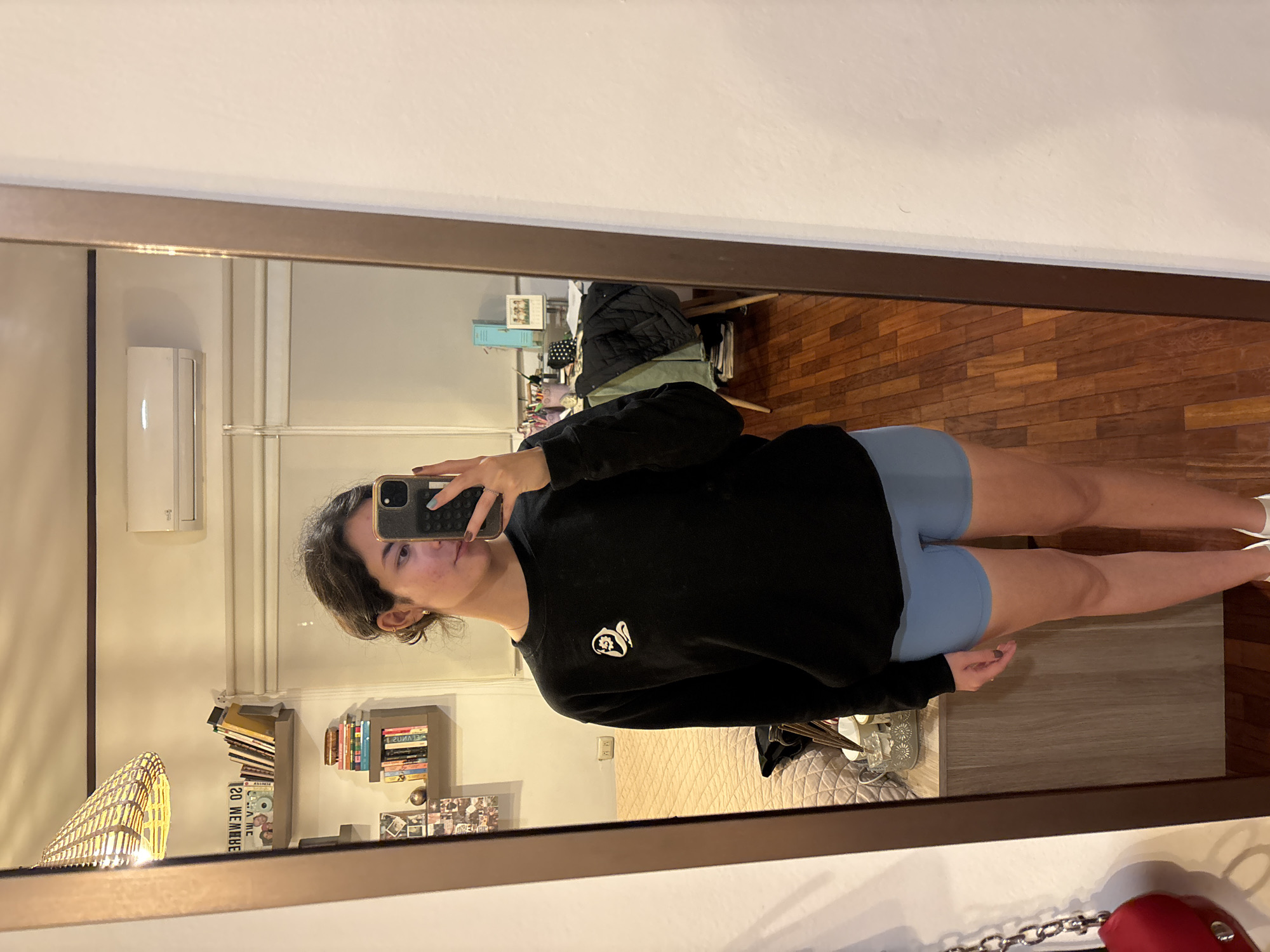 A young woman stands in front of a mirror, taking a selfie with her smartphone. She is wearing a black sweatshirt with a small white design on the left chest and light blue shorts. Her hair is dark and pulled back. The reflection in the mirror reveals a room with hardwood floors, bookshelves, a decorative lamp, and a wall-mounted air conditioning unit. The location is likely an indoor space, possibly an apartment, in a location presumed to be Puebla, Mexico.
