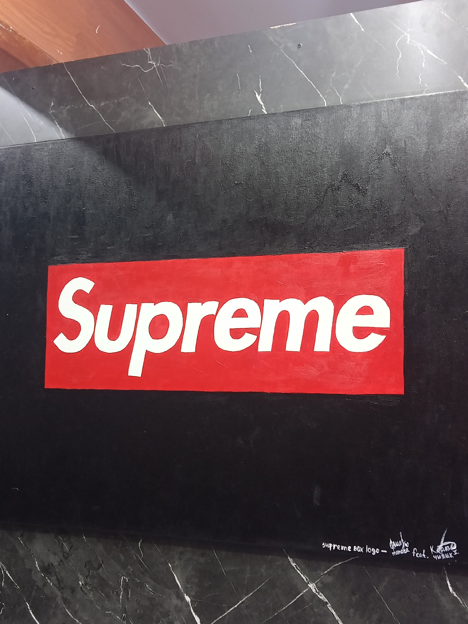 An oil painting on canvas depicts the iconic Supreme box logo. The logo, featuring white text on a red rectangular background, is centered on a black canvas. The black background has a textured appearance, possibly from the canvas weave or the application of paint. Below the logo, in the lower right corner of the canvas, there is faint white cursive handwriting, appearing to be a signature and possibly a title or dedication. The painting rests on a surface with a dark, grey, marble-like pattern, possibly a countertop or table. The lighting suggests an indoor environment.