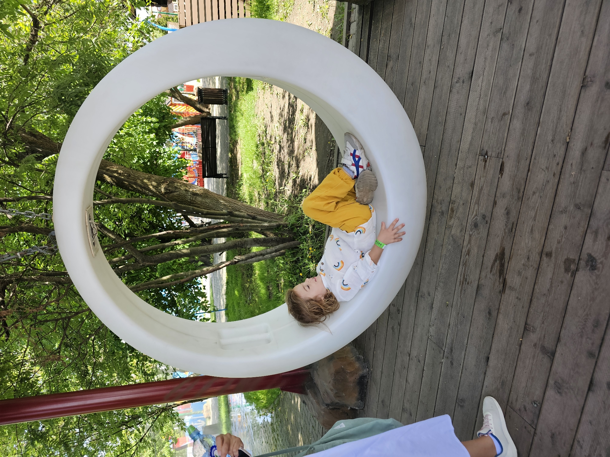 A child in yellow pants and a white shirt with rainbow designs lies inside a large, white circular structure. The structure appears to be a piece of playground equipment, situated on a wooden deck. Green trees and foliage surround the structure. In the background, playground equipment and benches are visible, suggesting a park setting. The person's left foot is visible wearing white trainers and pale blue trim. The image was taken in the city of Khimki, Russia.