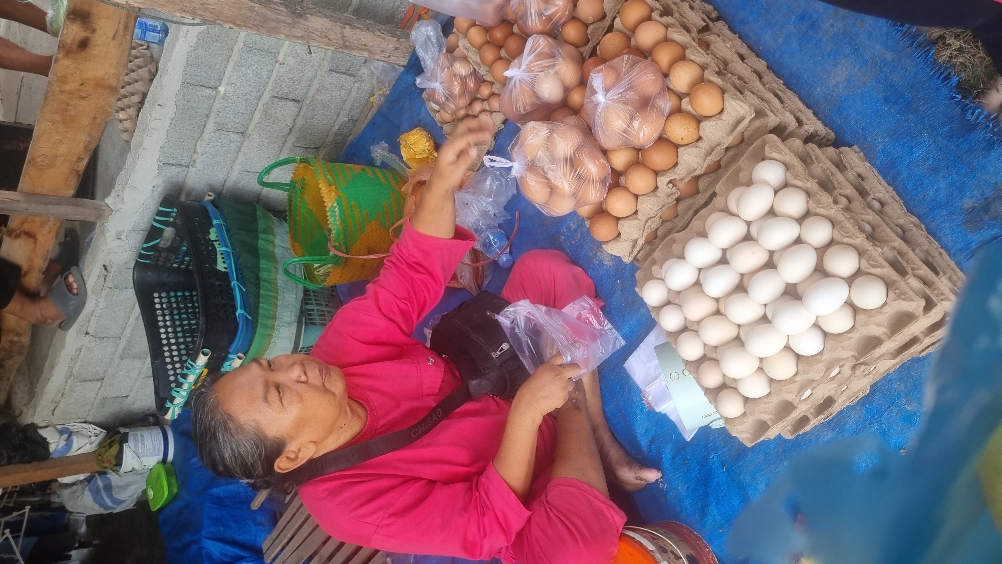 A woman wearing a bright pink shirt and pants sits cross-legged on a blue tarp. She has a black bag slung across her chest and appears to be interacting with a plastic bag in her hands. In front of her, arranged on the tarp, are stacks of egg cartons filled with eggs, both brown and white. To her right, more eggs are displayed in cartons and also in plastic bags. In the background, various baskets and containers are visible, along with portions of a brick wall and wooden structure. The scene suggests a market setting.