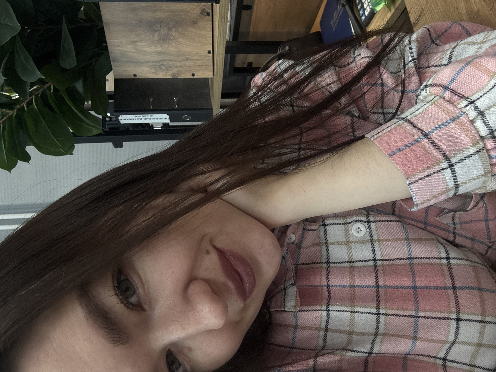 A close-up, rotated, and slightly blurry image shows a woman with long, dark hair wearing a pink and white plaid shirt. Her head is tilted, and her left cheek rests on her arm. Part of her face is visible, including her left eye, nose, and mouth with lipstick. Behind her are partially visible objects including a green leafy plant, a wooden box and part of a desk. The location in the city of Chaikovsky, Russia, cannot be determined from the image.