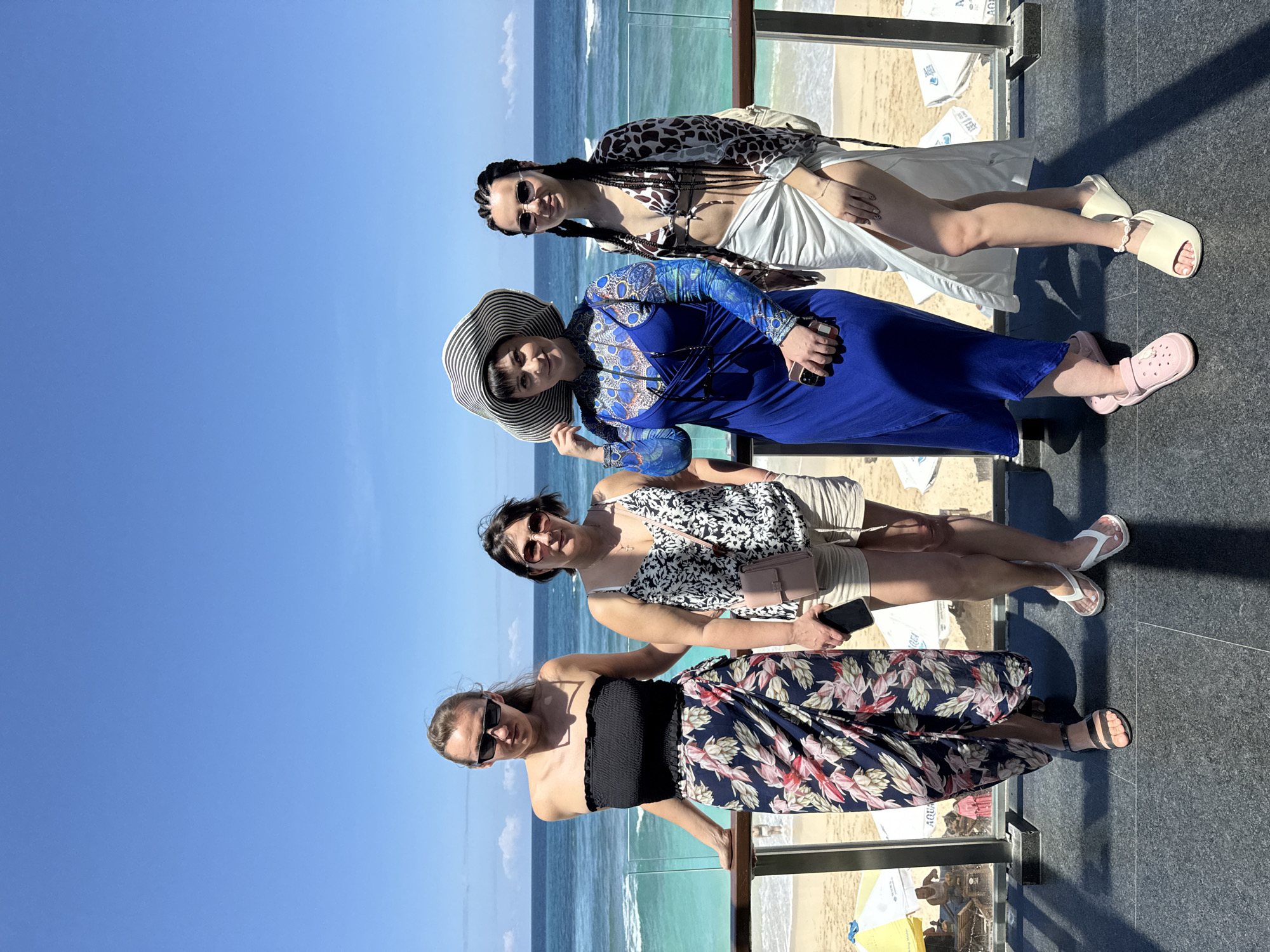 Four women are posing for a photo in the city of Kuta Selatan, Indonesia. The background features a beach, ocean, and blue sky. The women are standing on a dark grey stone platform with a glass railing.
From bottom to top, the first woman is wearing a black strapless top and a floral-patterned skirt. The second woman is wearing a black and white floral tank top and beige shorts, carrying a small purse and a smartphone. The third woman is wearing a long blue dress with a blue patterned long-sleeved top, and a striped floppy hat. The fourth woman is wearing a leopard print swimsuit top, a white skirt tied at the hip, and a matching animal print shawl. All the women are wearing sunglasses.