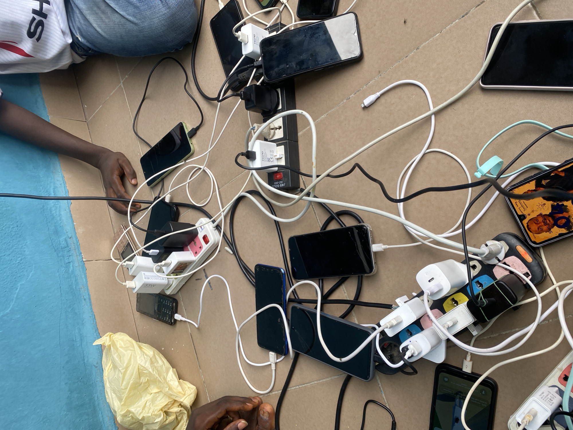 A group of multiple mobile phones are charging from multiple power strips, which are plugged into a wall socket. The phones and chargers are spread out on a tiled floor, with some cables also visible. The scene appears to be in an outdoor covered area, possibly a verandah or courtyard, with a blue wall in the background and a yellow plastic bag on the floor. A person's arm and shoulder are visible in the upper right corner of the frame, indicating a human presence, but no other individuals are clearly depicted. The overall impression is one of an informal charging station, likely in a residential or public space. The image was taken in Jalingo, Nigeria.