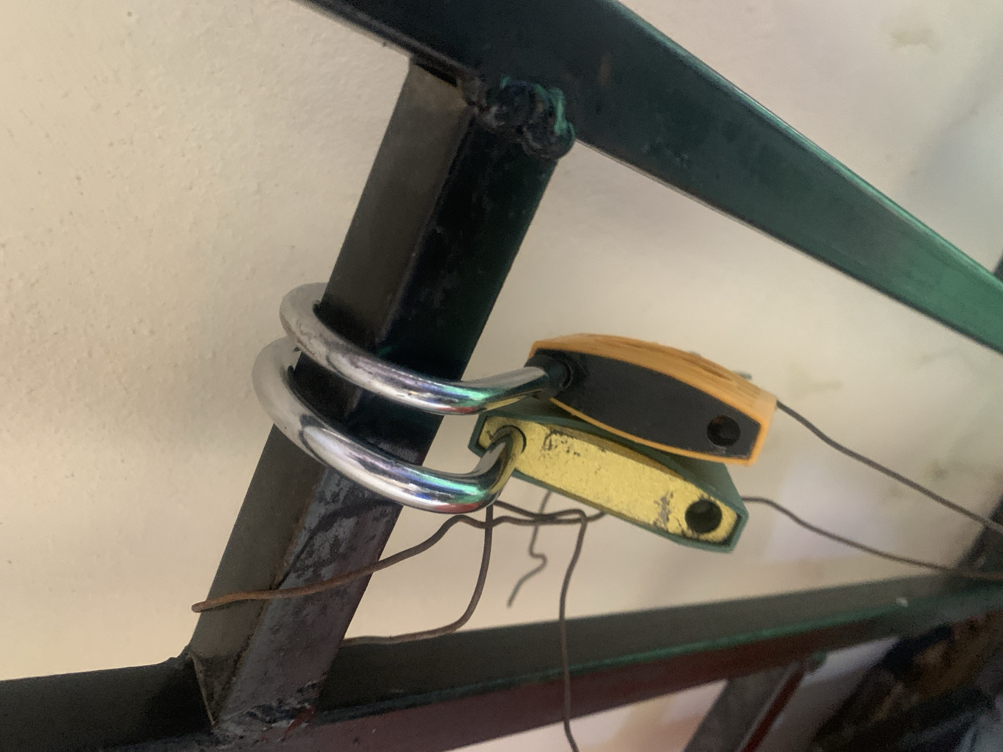 The image shows two padlocks, one yellow and one orange and black, hanging from a black metal structure. Both padlocks have their shackles engaged through a loop of the metal structure. Thin, dark wires are also visible, attached to the metal structure and hanging loosely. The background is a textured, light-colored wall. The scene appears to be outdoors or in a space with a textured wall. The image does not provide any information to determine the location in Jauro Yinu, Nigeria.