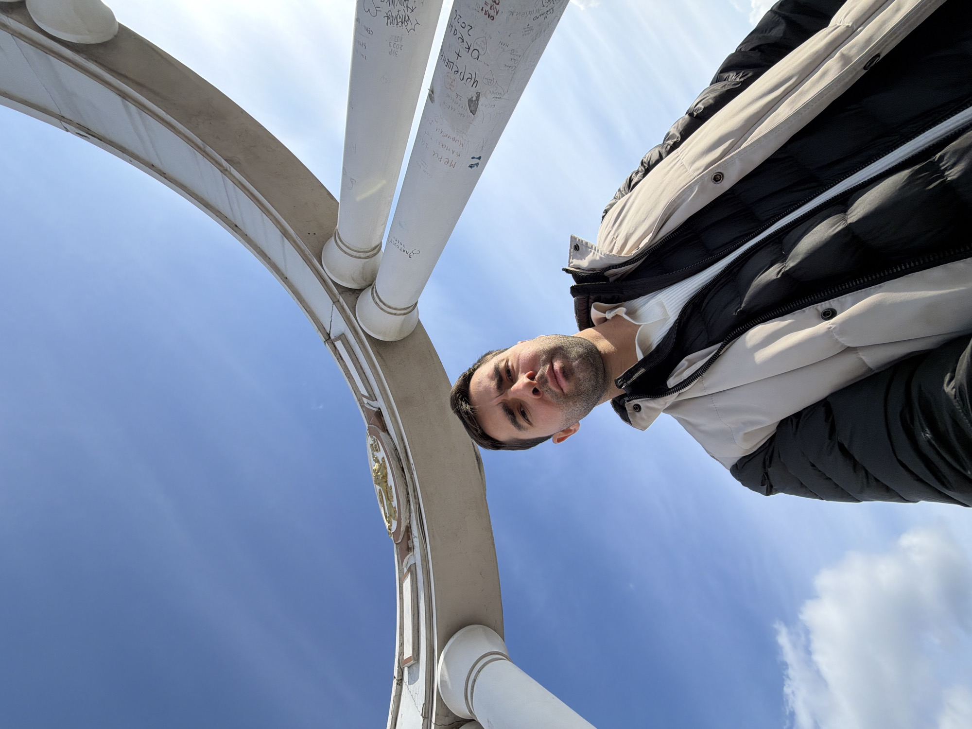 The image is a low-angle selfie taken outdoors, with the subject being a man wearing a black and beige puffer jacket and a white shirt. The background features a partially visible white arched structure with graffiti, set against a blue sky with some clouds. The structure appears to be part of an architectural monument. The subject's face is partially visible in the frame. Graffiti is visible on the pillars of the structure. Based on the information embedded in the image, the location is Krasnoyarsk, Russia.