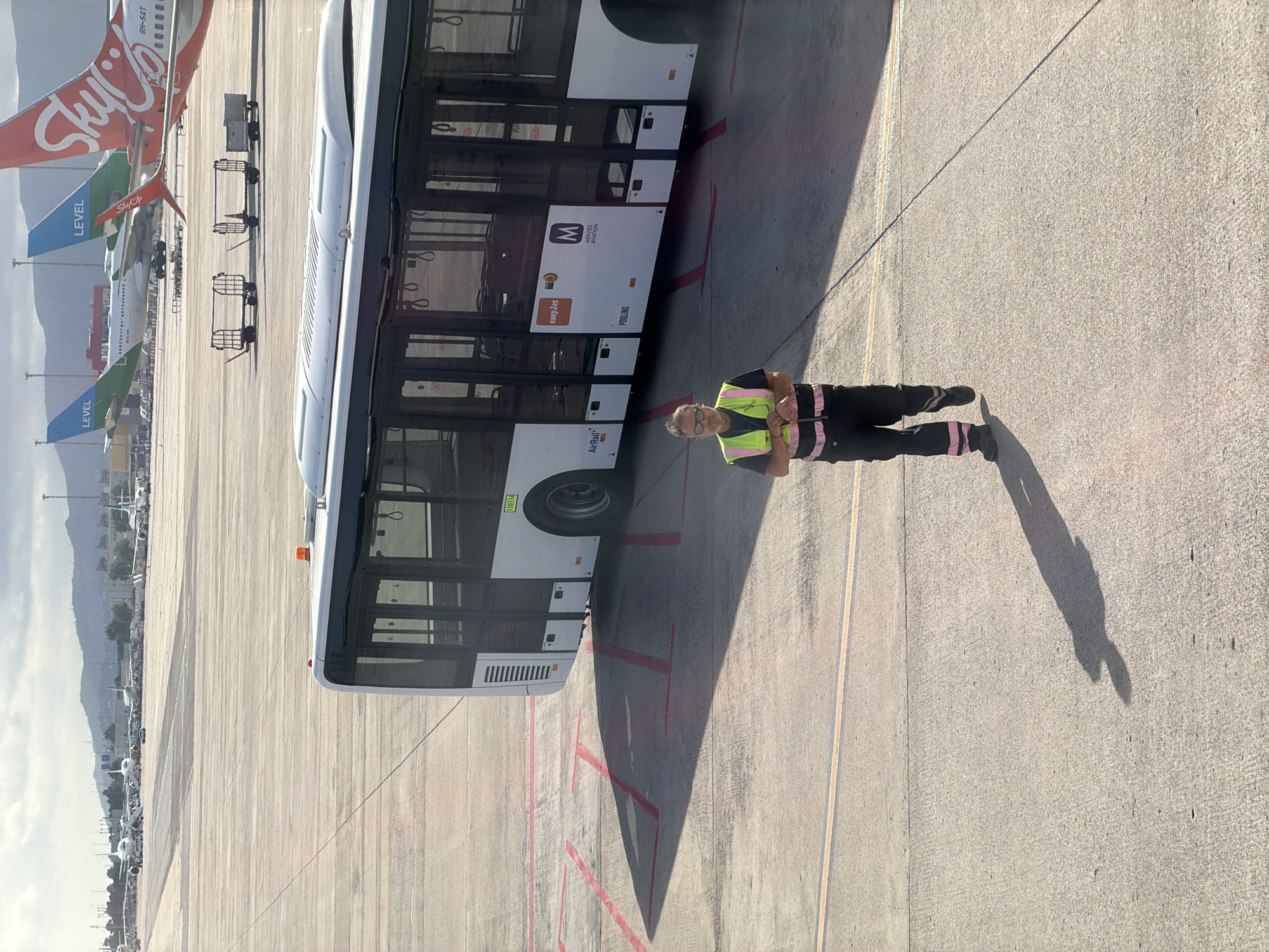 A man in a high-visibility vest and dark pants stands on an airport tarmac next to a white bus with tinted windows. The bus has "M MENZIES AVIATION" and "easyJet" written on its side. The man appears to be looking towards the camera, his hands clasped in front of him. His shadow stretches across the tarmac, indicating a sunny day. In the background, several airplanes are parked, including aircraft with "LEVEL" and "SkyUp" branding. The overall setting is an airport, likely in Barcelona, Spain, given the provided location context. The image captures a moment of a worker's presence at the airport, with the bus and planes suggesting ongoing operations or passenger transport. The lighting suggests it is daytime, possibly late afternoon due to the long shadows.