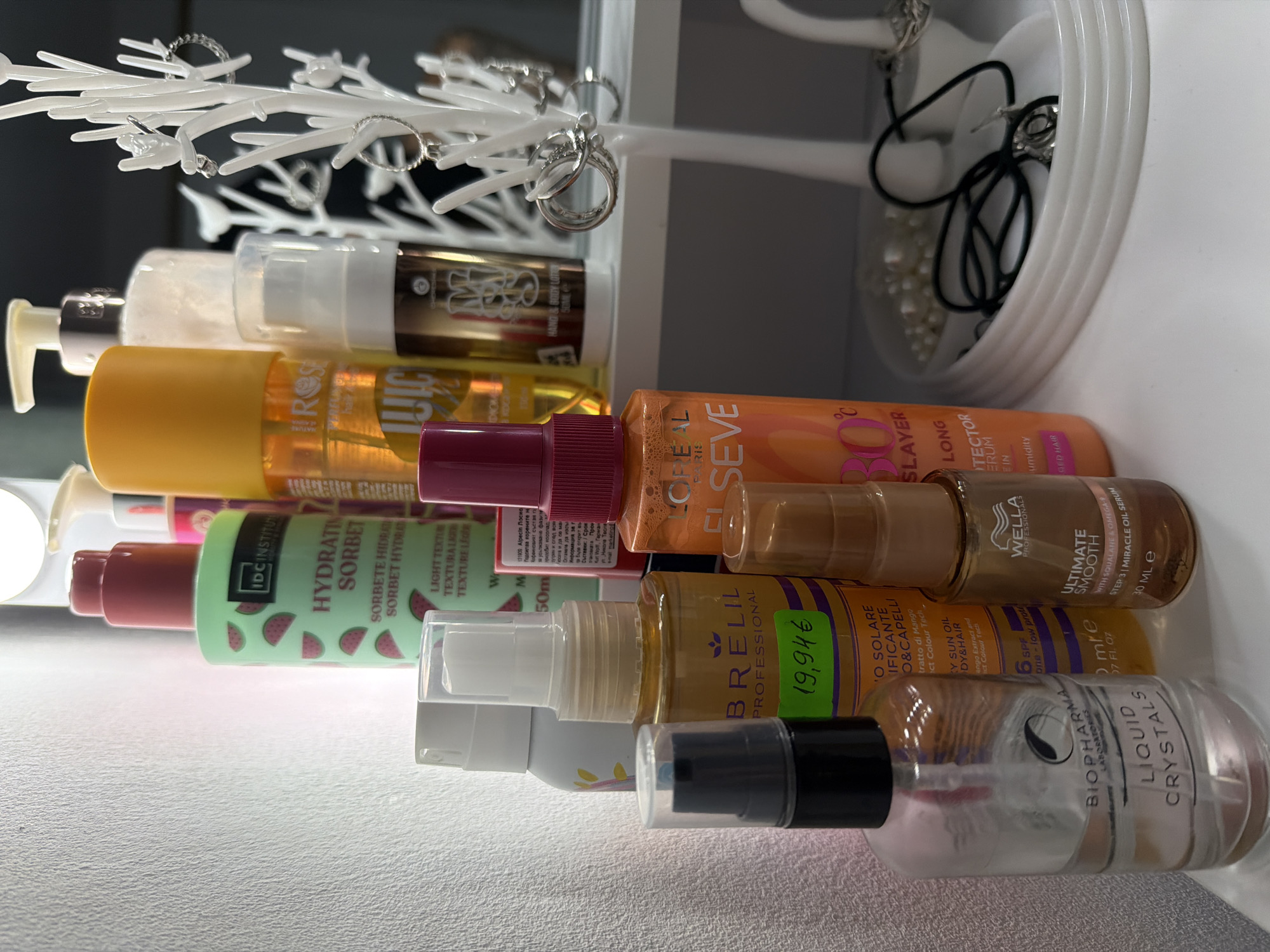 The image depicts a collection of cosmetic products and jewelry arranged on a white surface, possibly a vanity or shelf, set against a textured, pale wall. The items are primarily bottles and containers of various sizes and colors, suggesting a personal grooming or beauty-focused setting. Among the visible items are several bottles of hair products, including one with a green label featuring watermelons and the text "HYDRATING SORBET," and another orange bottle with "L'OREAL" branding. There's also a clear bottle labeled "BIOPHARMA LIQUID CRYSTALS." Jewelry, including rings and necklaces, is displayed on a white tree-like stand and in a white tray filled with pearls. The lighting in the scene is bright, with some reflections visible on the product surfaces. The overall impression is one of organized personal care items. This scene is located in the city of Plovdiv, Bulgaria.