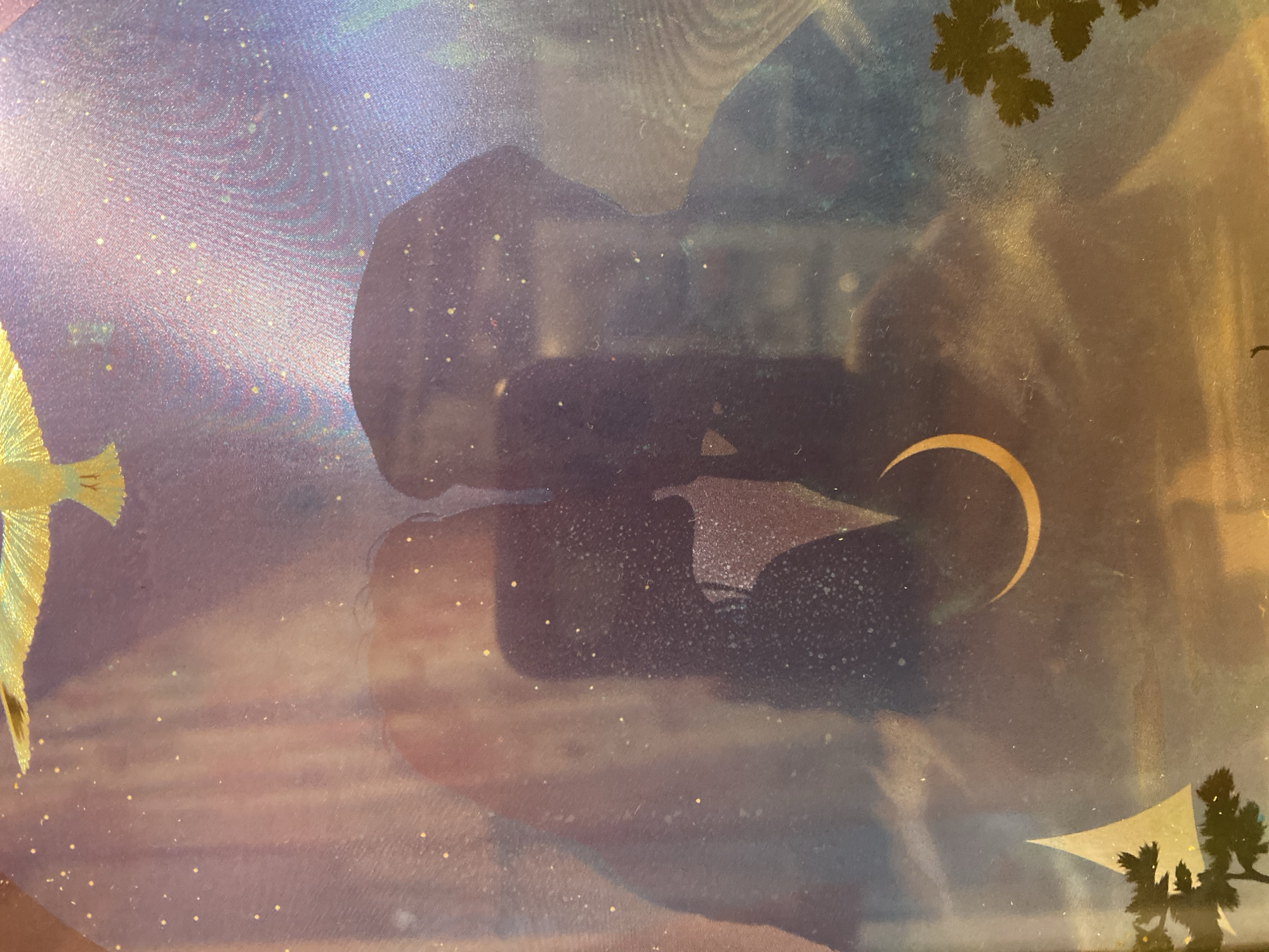 This is a close-up, abstract photographic image with a textured, possibly fabric, background. The dominant colors are shades of purple, blue, and yellow, creating a dreamy and ethereal atmosphere. There are small, scattered yellow specks that resemble stars or glitter.
On the left side of the image, a golden object with radiating lines appears to be a stylized bird or wing. In the center, there is a dark, amorphous shape with lighter speckled areas. Emerging from this shape, a thin, golden crescent arc is visible, suggestive of a moon or celestial body.
In the upper right corner, dark green, leaf-like shapes are silhouetted against a yellow-orange gradient, hinting at foliage. In the lower right, a white, triangular shape is also silhouetted, possibly part of a sail or abstract form, with more dark green foliage beneath it.
The overall impression is one of a surreal or fantastical scene, perhaps a dreamscape or a decorative artwork. There is no discernible activity or event taking place. The lighting appears diffused, and there are no clear indicators of time of day, weather, or a specific location within Novaya Usman', Russia, beyond what might be inferred from artistic motifs. No visible text is present in the image. The texture of the surface suggests it might be a printed fabric, a painted canvas, or a similarly textured material.