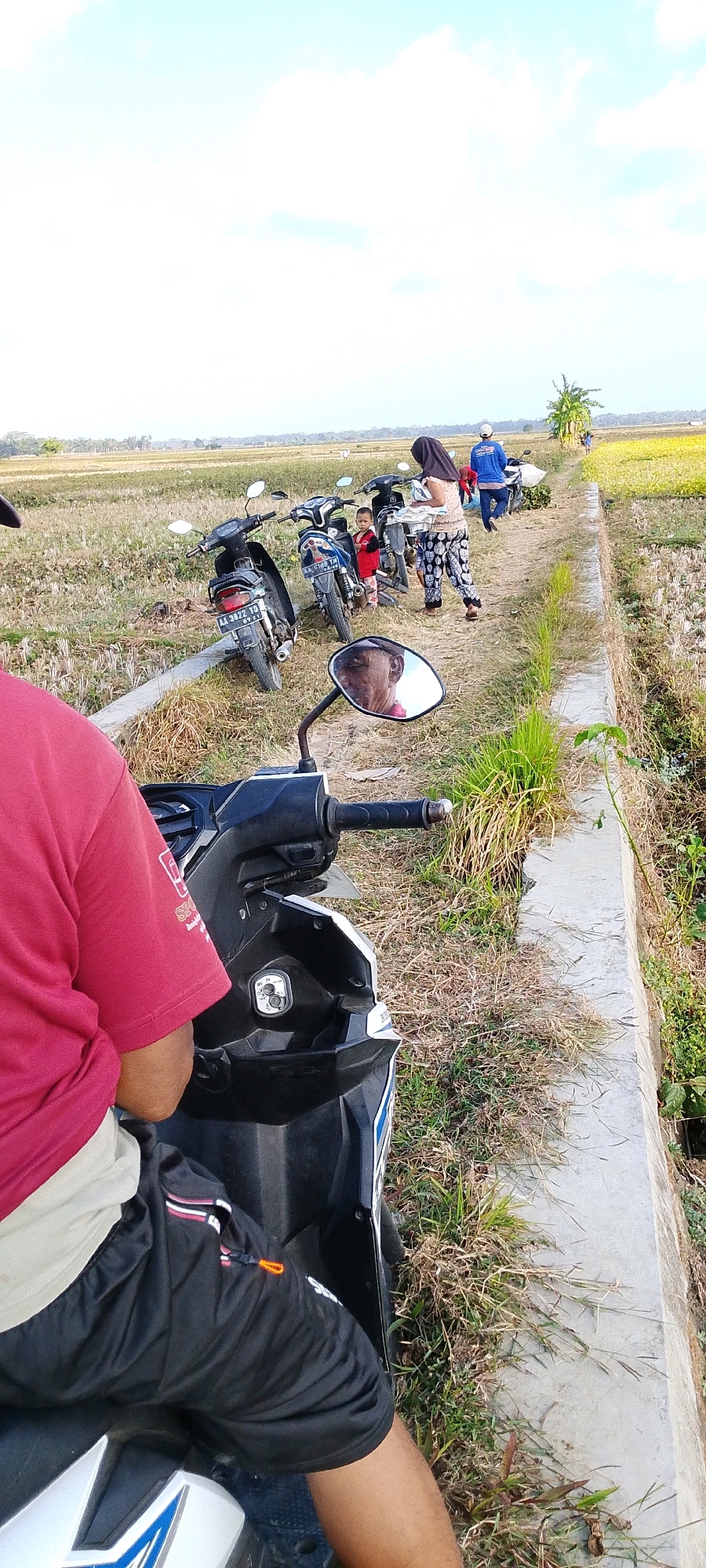 The image depicts a rural scene in Geblug, Indonesia, with a focus on a group of people and motorcycles in a dry, grassy field adjacent to a concrete irrigation channel. In the foreground, the back of a person wearing a red shirt and black shorts is visible, seated on a black motorcycle. The motorcycle's handlebars, front panel, and a rearview mirror are prominent. The mirror reflects the face of a person wearing a red shirt.
In the middle ground, several motorcycles are parked in a line. A woman wearing a hijab and patterned pants stands near one of the motorcycles, holding what appears to be a stack of white clothing. A small child in a red shirt stands beside her. Further down the line of motorcycles, another person in a blue shirt and a white cap walks away from the camera, carrying a white bag.
The environment is characterized by dry, straw-colored grass and some patches of green vegetation, suggesting a dry season. The concrete channel runs along the right side of the frame, with small plants growing on its edge. In the background, a vast, flat landscape stretches out, likely agricultural fields, under a bright, cloudy sky. A lone banana tree stands to the right of the motorcycles. The overall impression is one of a casual gathering or stop in a rural Indonesian setting.
