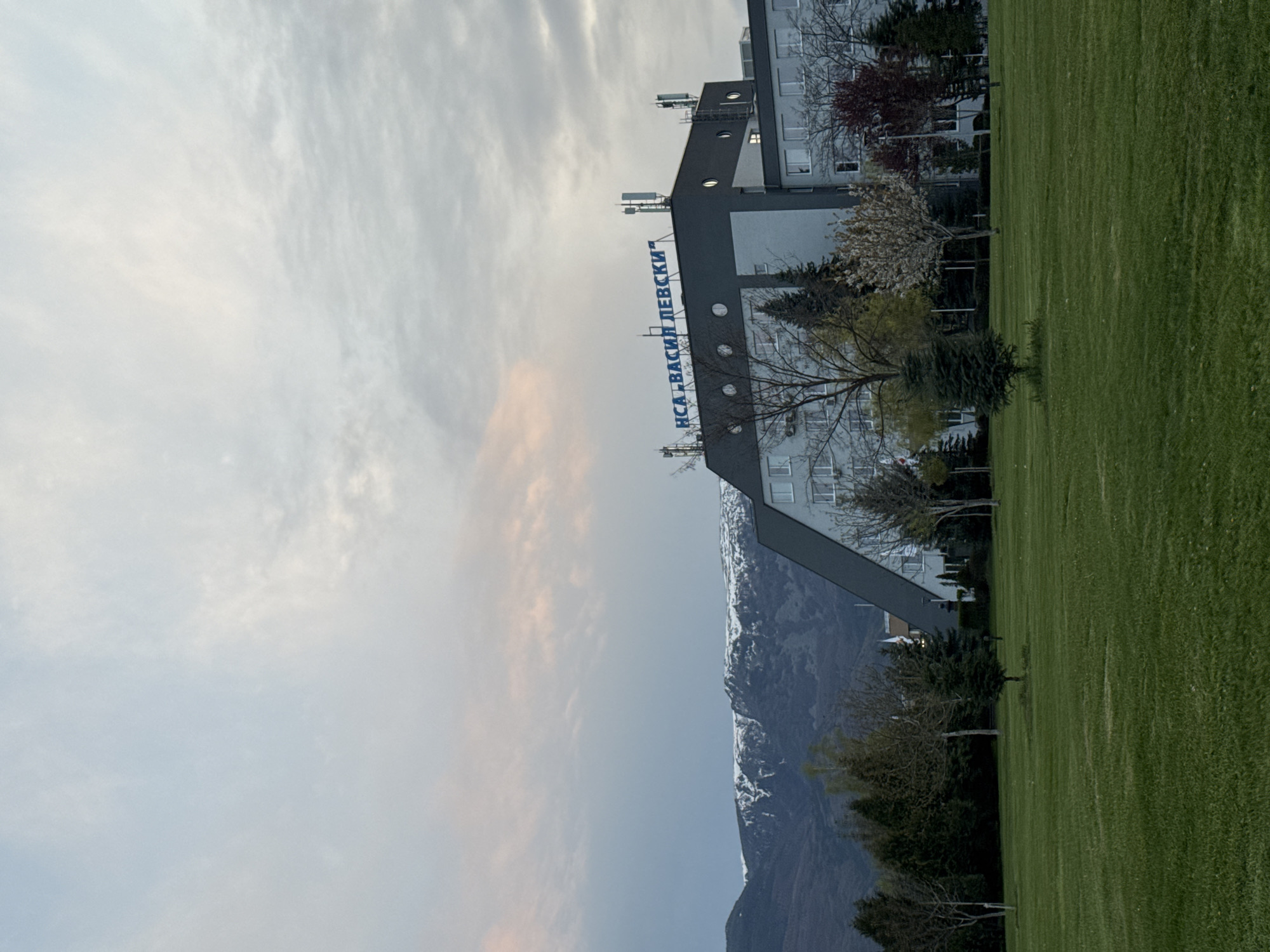 The image captures a panoramic view of a modern building, likely an institutional complex, set against a backdrop of mountains and a vast green landscape. The scene appears to be set in Sofia, Bulgaria, during the late afternoon or early evening hours, suggested by the soft, diffused light and the pastel colors in the sky.
In the mid-ground, a multi-story building with a distinctive architectural style occupies the left side of the frame. It features a dark grey roofline and upper section, contrasting with lighter grey or white walls, and multiple rectangular windows. Prominently displayed on the building's facade are large blue Cyrillic letters spelling out "НСА „ВАСИЛ ЛЕВСКИ“" (NSA "Vasil Levski"), indicating it is the National Sports Academy "Vasil Levski." Several antennae or communication structures are visible on its roof.
The foreground and extends to the right is covered by a meticulously maintained, vibrant green lawn or sports field. Various trees are scattered across the scene; some are bare-branched, others show early spring blossoms or budding green leaves, and several are dark evergreen trees.
Behind the building and trees, a majestic range of mountains rises, with visible patches of snow near their summits. The sky above is partly cloudy, displaying soft gradients of light blue, grey, and subtle pinkish-orange hues. The overall atmosphere is tranquil, with no people or specific activities visible.