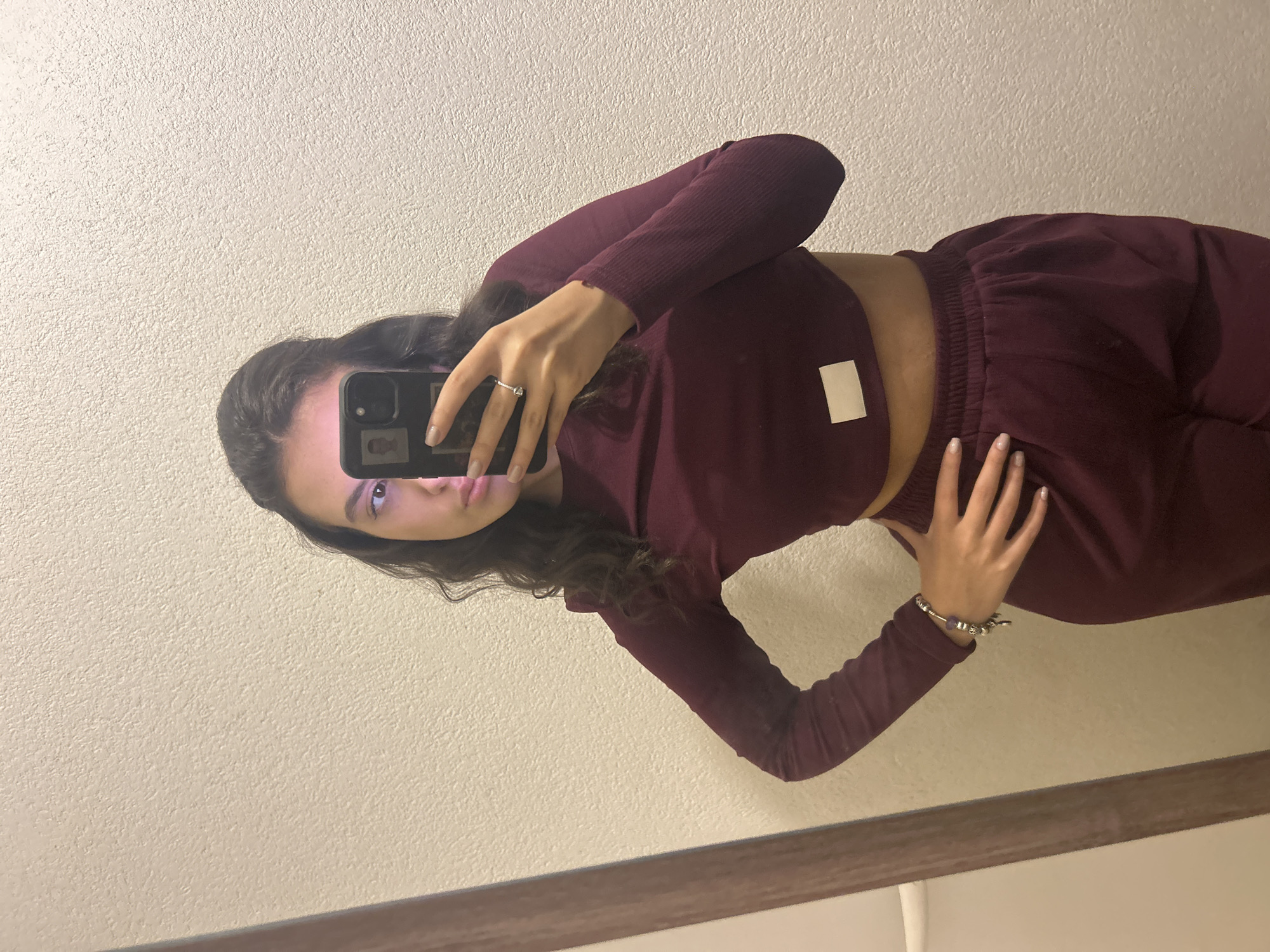 A woman takes a selfie in a mirror. She is wearing a matching burgundy-colored cropped long-sleeve shirt and sweatpants. She holds a black smartphone in her right hand, partially obscuring her face. She wears a ring on her left ring finger and a bracelet on her left wrist. The background consists of a light-colored wall and the edge of a mirror frame. The location is Unknown, Unknown.