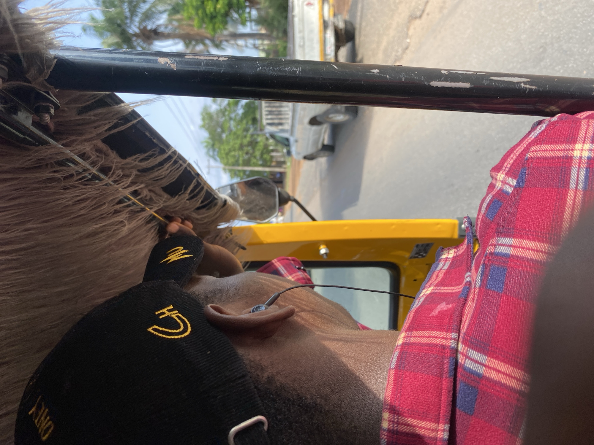 The image shows the interior of a yellow auto-rickshaw in Jalingo, Nigeria. The back of a person wearing a red and blue plaid shirt and a black cap with yellow embroidery is visible. The person has earphones in their ears. Outside the auto-rickshaw, a white pickup truck is driving on a paved road, with trees and palm trees lining the sides of the road under a bright, clear sky. The scene appears to be a sunny day.