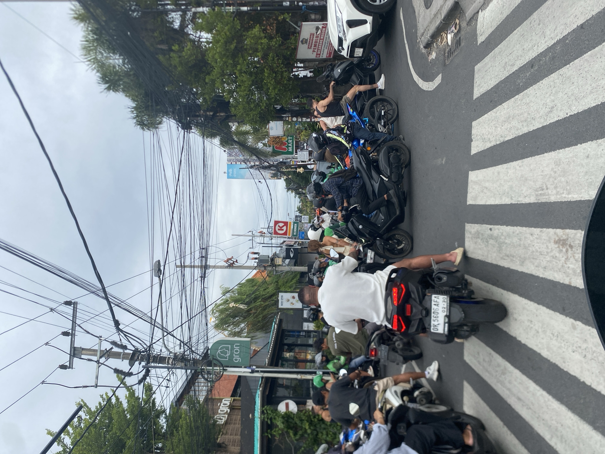 The image depicts a crowded street scene in North Kuta, Indonesia, with numerous motorcycles and a few cars navigating a congested intersection. The sky is overcast, suggesting possibly humid or rainy weather. Overhead, a complex network of electrical wires is prominently visible, strung between utility poles.
In the foreground, several individuals on motorcycles are stopped at a crosswalk. One person in a white t-shirt is seated on a black motorcycle, facing away from the camera, their rear brake lights illuminated. To the left, another motorcycle rider in a dark shirt is also stopped.
Further into the scene, more motorcycles are visible, some with passengers. A white SUV is positioned on the right side of the road, partially obscuring other vehicles. Various shop fronts and signage are present along the street, including what appears to be a "Gron" establishment with a green logo. A "K Mart" sign is also discernible.
The overall impression is one of a busy urban environment where motorbikes are the primary mode of transportation, leading to significant traffic density. The infrastructure includes utility poles with abundant wiring and paved roadways marked with pedestrian crossings.