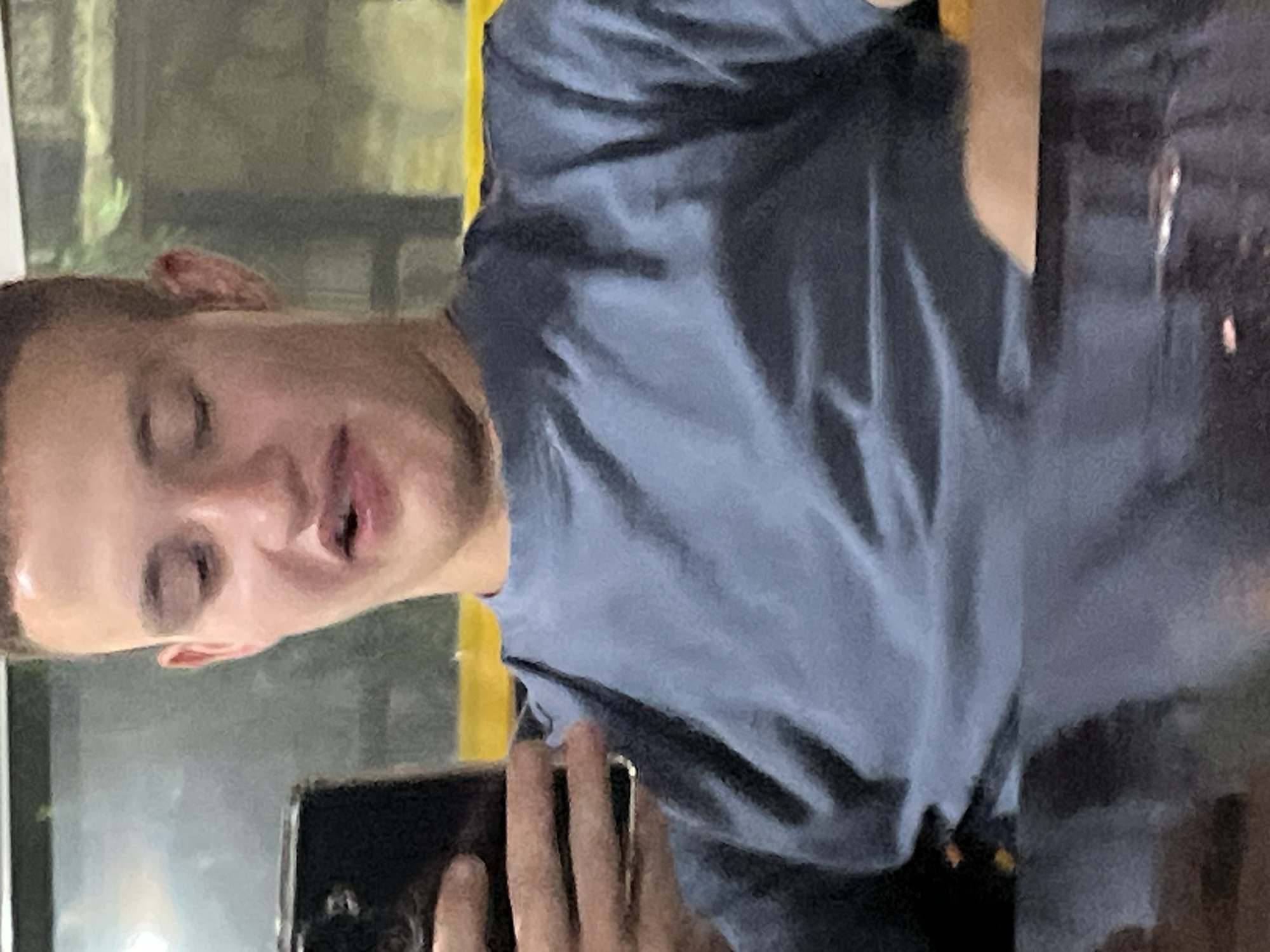 The image displays a young adult male indoors, seated at a dark, reflective table within what appears to be a public establishment in Basarbovo, Bulgaria. The individual has short, dark hair and light skin. His eyes are partially closed or squinted, and his mouth is open with the tongue visible, slightly extended, and curved, forming a distinct grimace or expression of distaste. He is wearing a plain, light blue, short-sleeved crew-neck t-shirt. His left hand holds a black smartphone vertically; the phone's screen is not visible. Through a large window in the background, blurred green foliage and a prominent horizontal yellow band are discernible, indicating an exterior view. The foreground consists of the dark, reflective surface of the table.