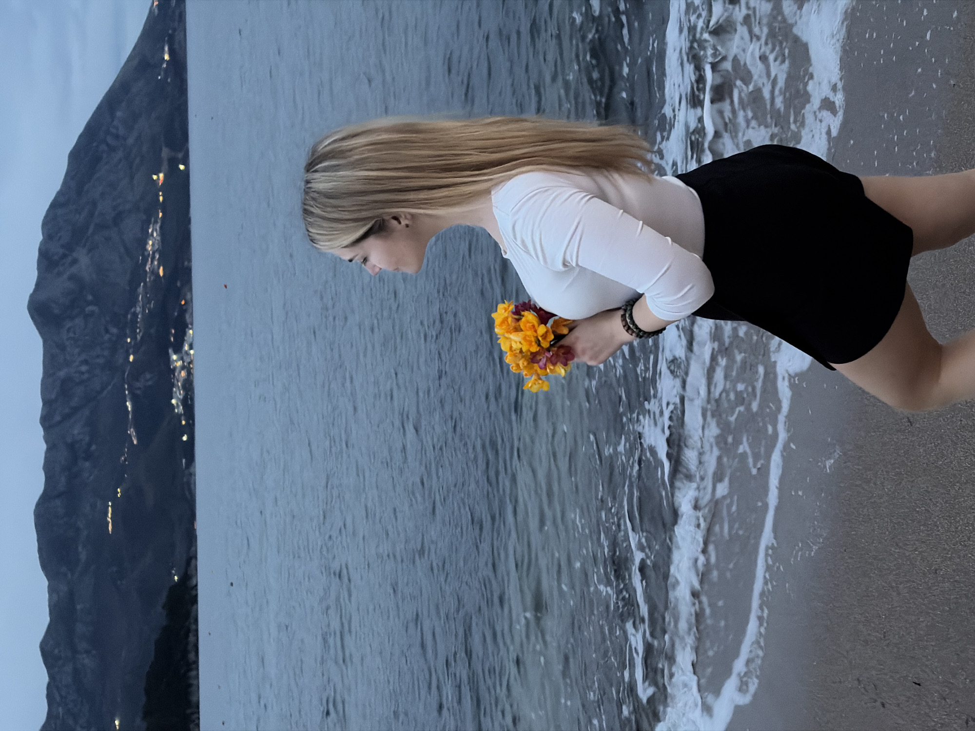 A young woman with blonde hair stands on a sandy beach, holding a bouquet of yellow and maroon flowers. She wears a white long-sleeved shirt and a black skirt. The sea's waves are washing onto the shore beside her. In the background, a dark, mountainous landscape is visible with scattered lights, indicating a city or town nestled against the mountain. The sky is overcast, suggesting either dawn or dusk. This is likely the Budva Riviera area.