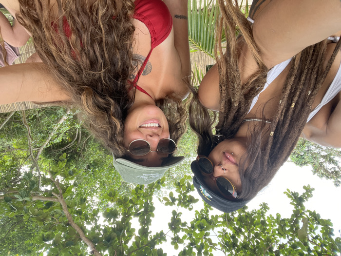 A selfie captures two women positioned outdoors in Baan Sri Thanu, Thailand. The woman on the left has long dreadlocks with golden beads, wears a black cap, oval sunglasses, a silver choker, and a white bikini top. She exhibits multiple facial piercings, including a septum ring, a nostril ring, and a labret piercing, and looks towards the camera with a subtle expression. Her right arm is extended, indicating she is holding the camera. The woman on the right has long, wavy brown hair, wears an olive green baseball cap, round sunglasses, and a red bikini top. She displays a wide smile, a visible tan line on her chest, and a tattoo on her left upper chest. The background features dense green foliage from trees and branches overhead, suggesting a tropical environment. A natural bamboo-like screen or fence is visible behind the subjects, and a partial view of another person is present in the far right background. The sky is bright but obscured by the tree canopy.