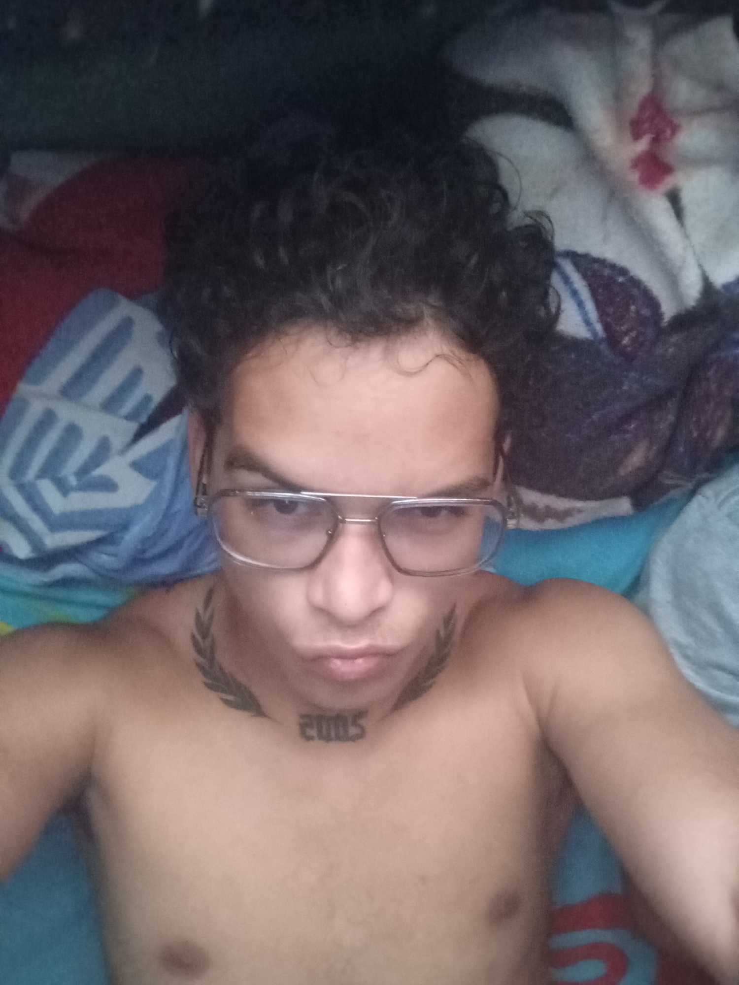 A shirtless person is lying on a bed. The person has curly, dark hair and wears eyeglasses. The person has tattoos on their neck: one appears to be a laurel wreath, and the other reads "2005". The bed has several blankets or sheets with different patterns and colors, including blue, red, and white. The location is not discernible from the image.